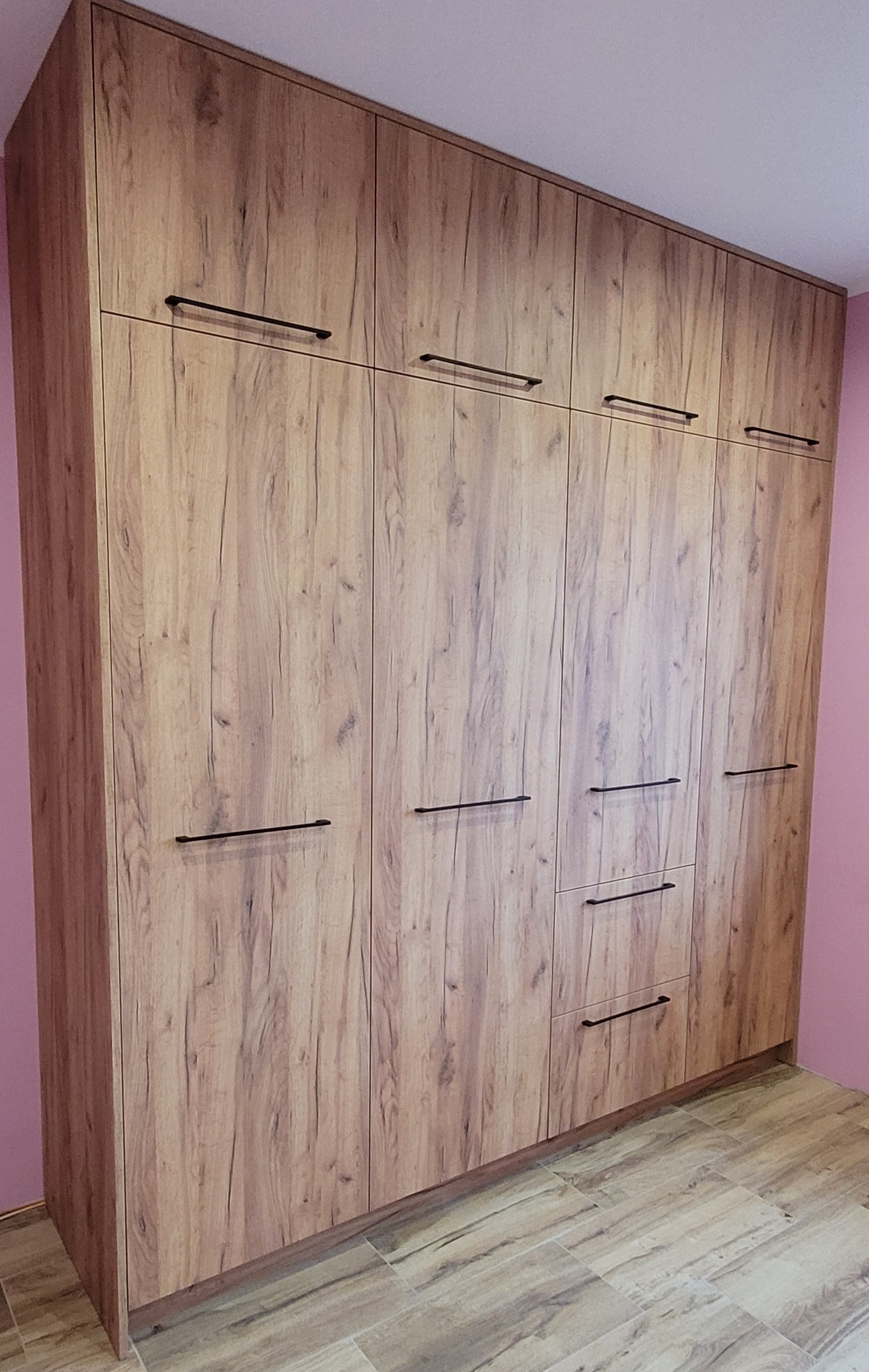 Duża, drewnopodobna szafa wnękowa z czarnymi, minimalistycznymi uchwytami, sięgająca sufitu w pokoju o różowych ścianach.