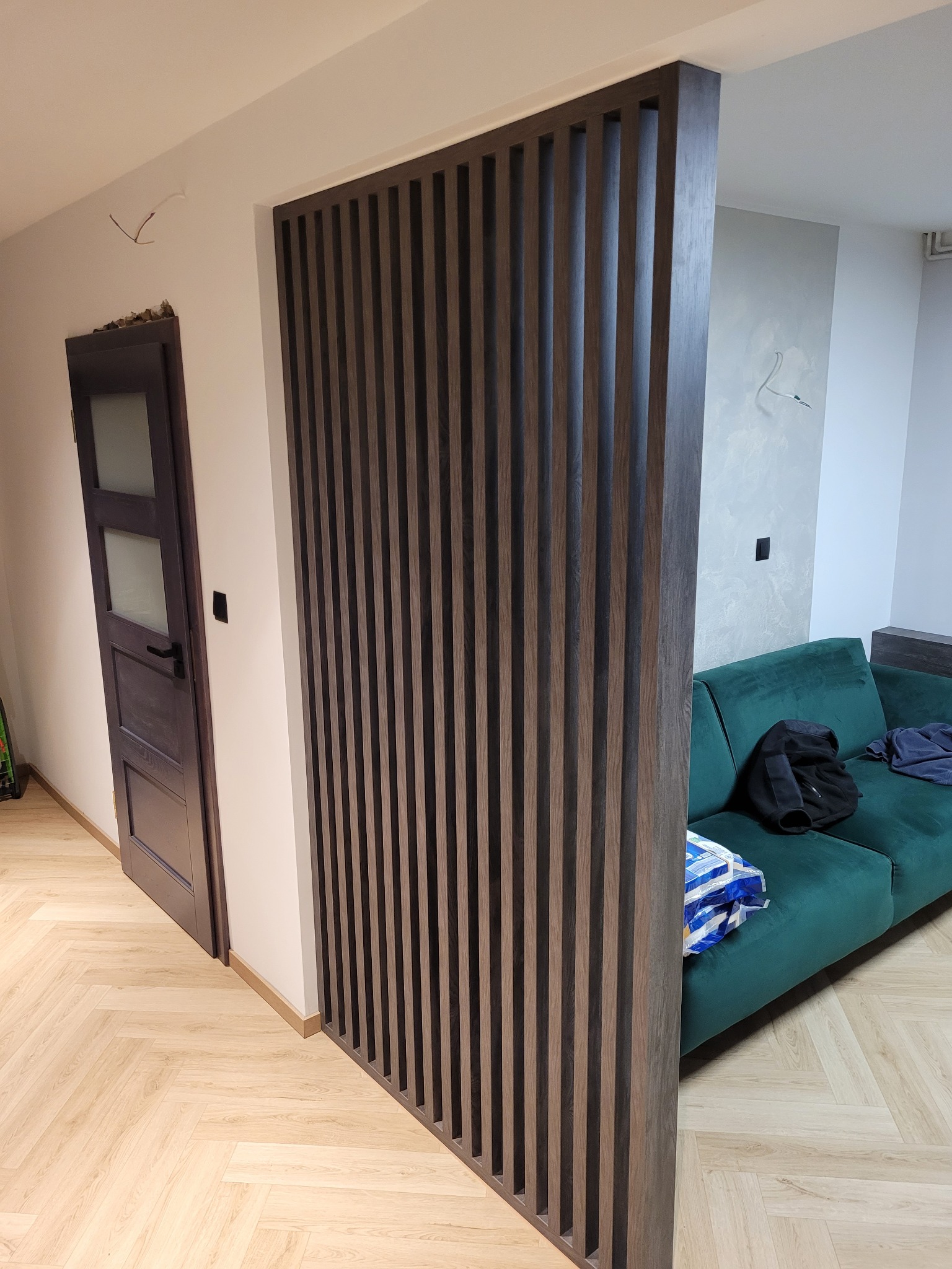 Drewniana ścianka działowa z pionowych listew w kolorze ciemnego drewna, oddzielająca przestrzeń salonu z zieloną sofą od przedpokoju z drewnianą podłogą w jodełkę i drzwiami.