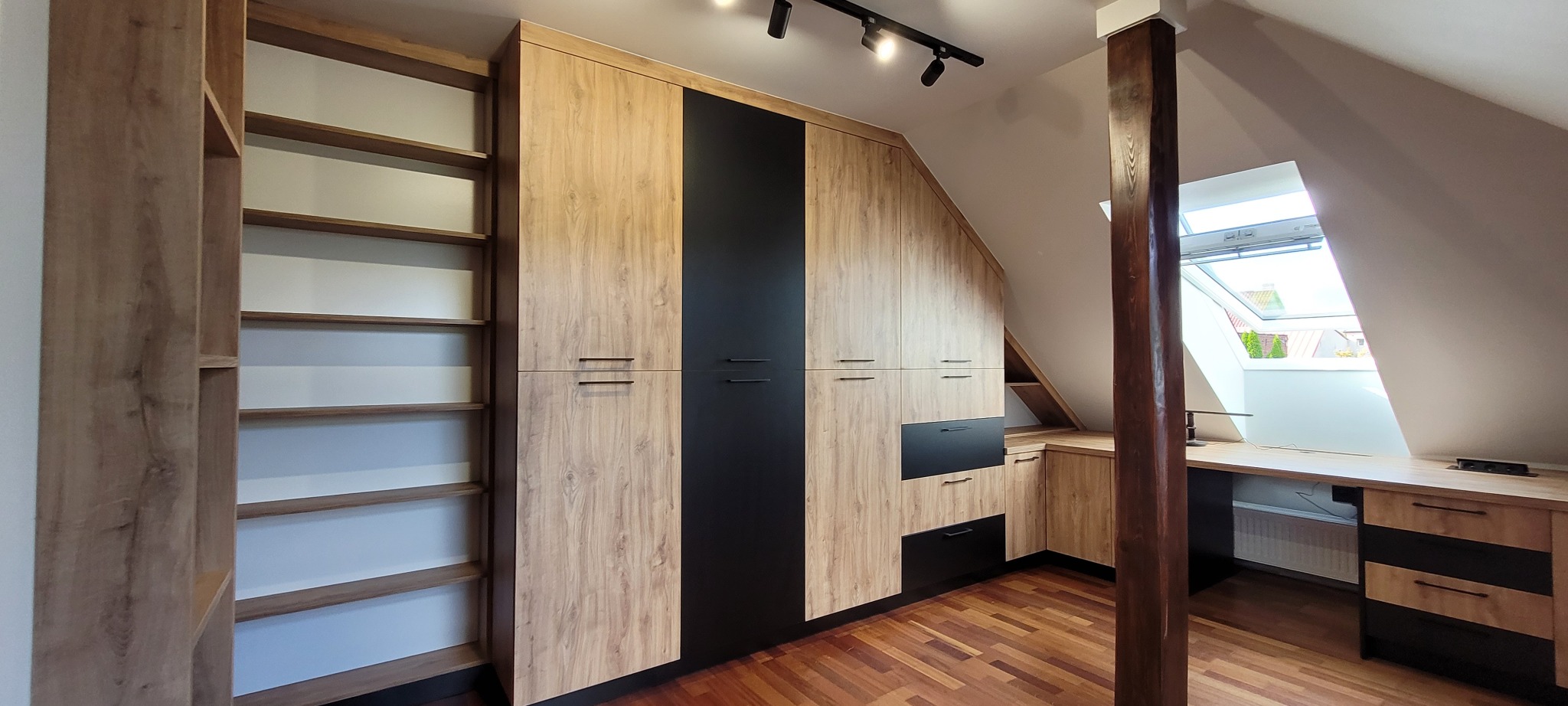 Adaptacja poddasza: zabudowa meblowa z szafami w kolorze drewna i czerni, półki regałowe, biurko pod oknem dachowym, drewniana podłoga parkietowa.