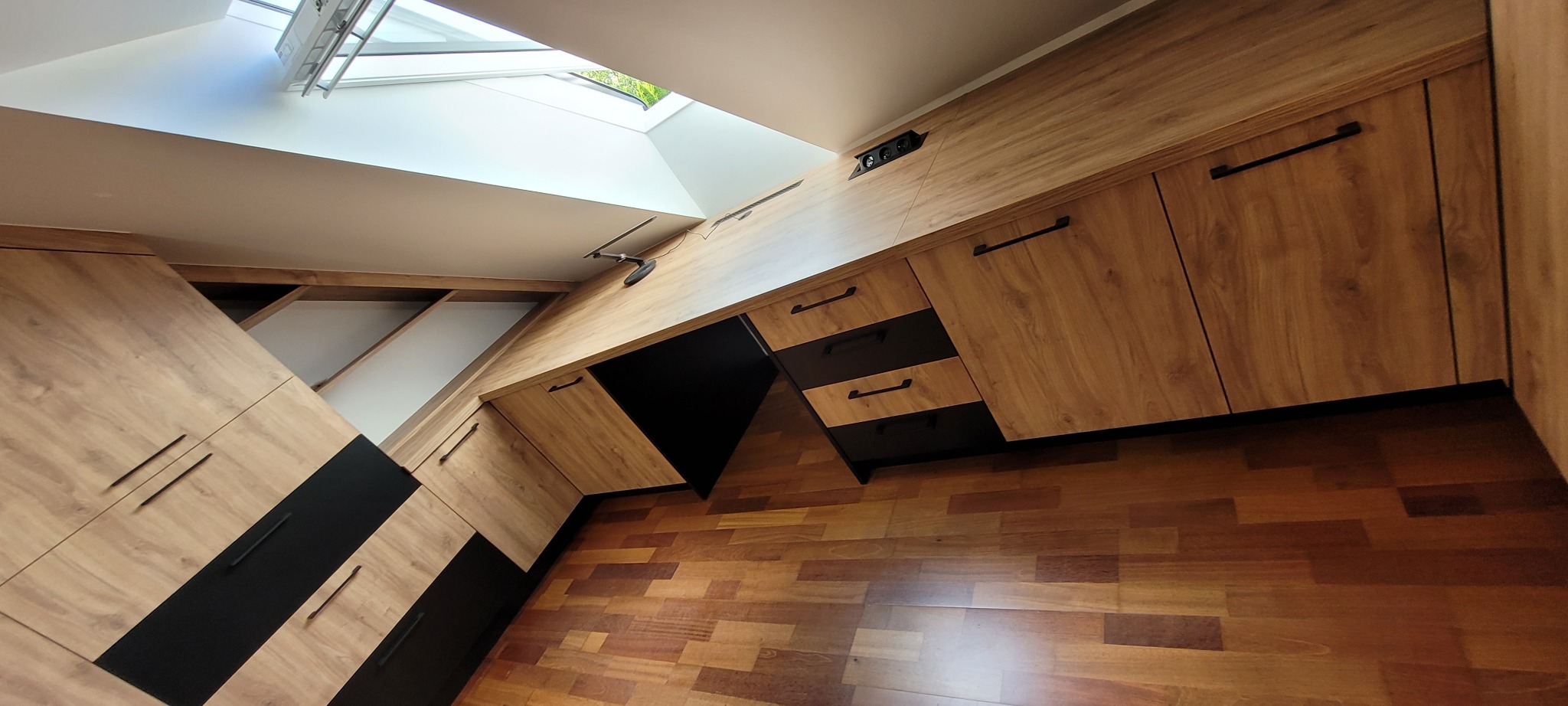 Funkcjonalna zabudowa meblowa z drewnopodobnymi frontami i czarnymi akcentami, wkomponowana w pomieszczenie z oknem dachowym, widoczna pod kątem.