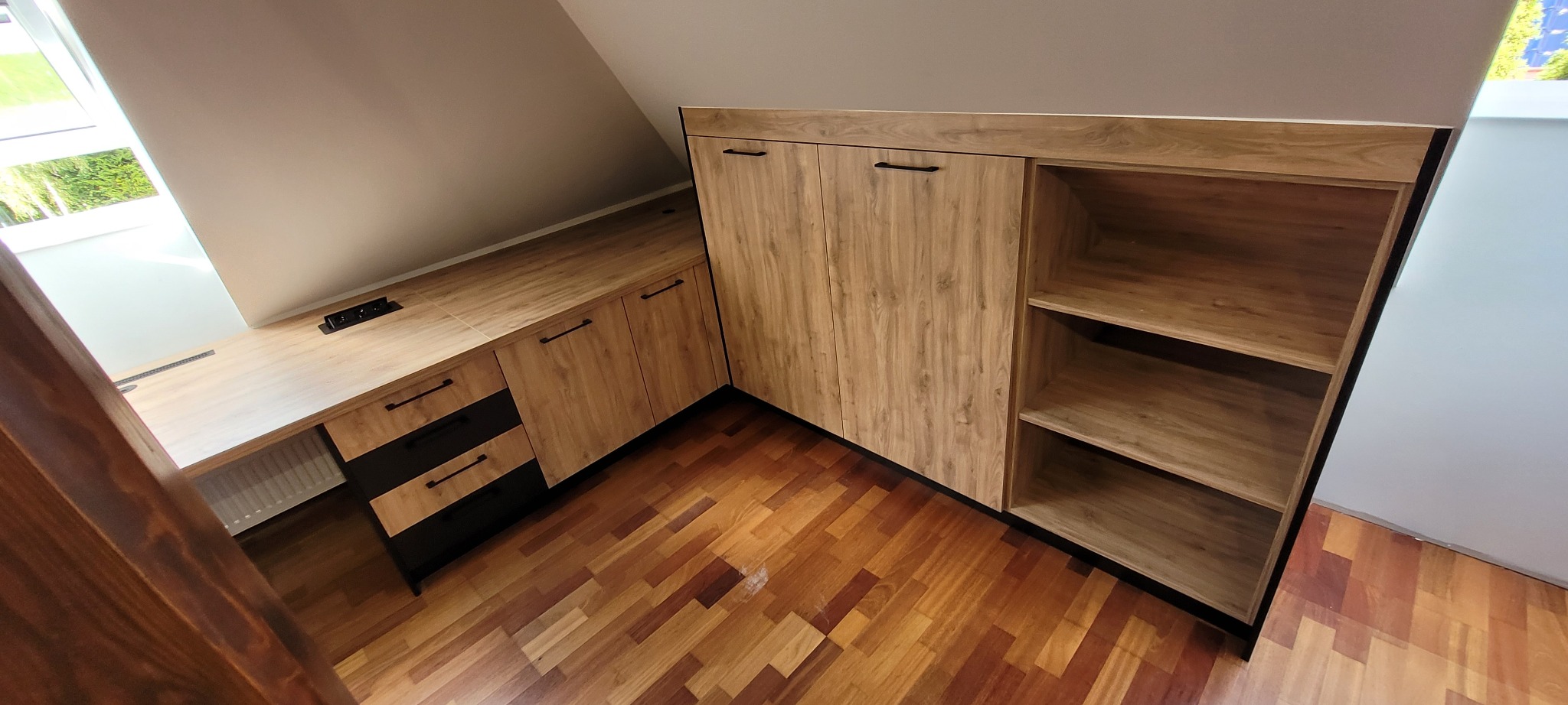 Zabudowa pod skosem z drewnianymi frontami i czarnymi detalami, obejmująca szafę z półkami i biurko z szufladami, widoczny parkiet na podłodze.
