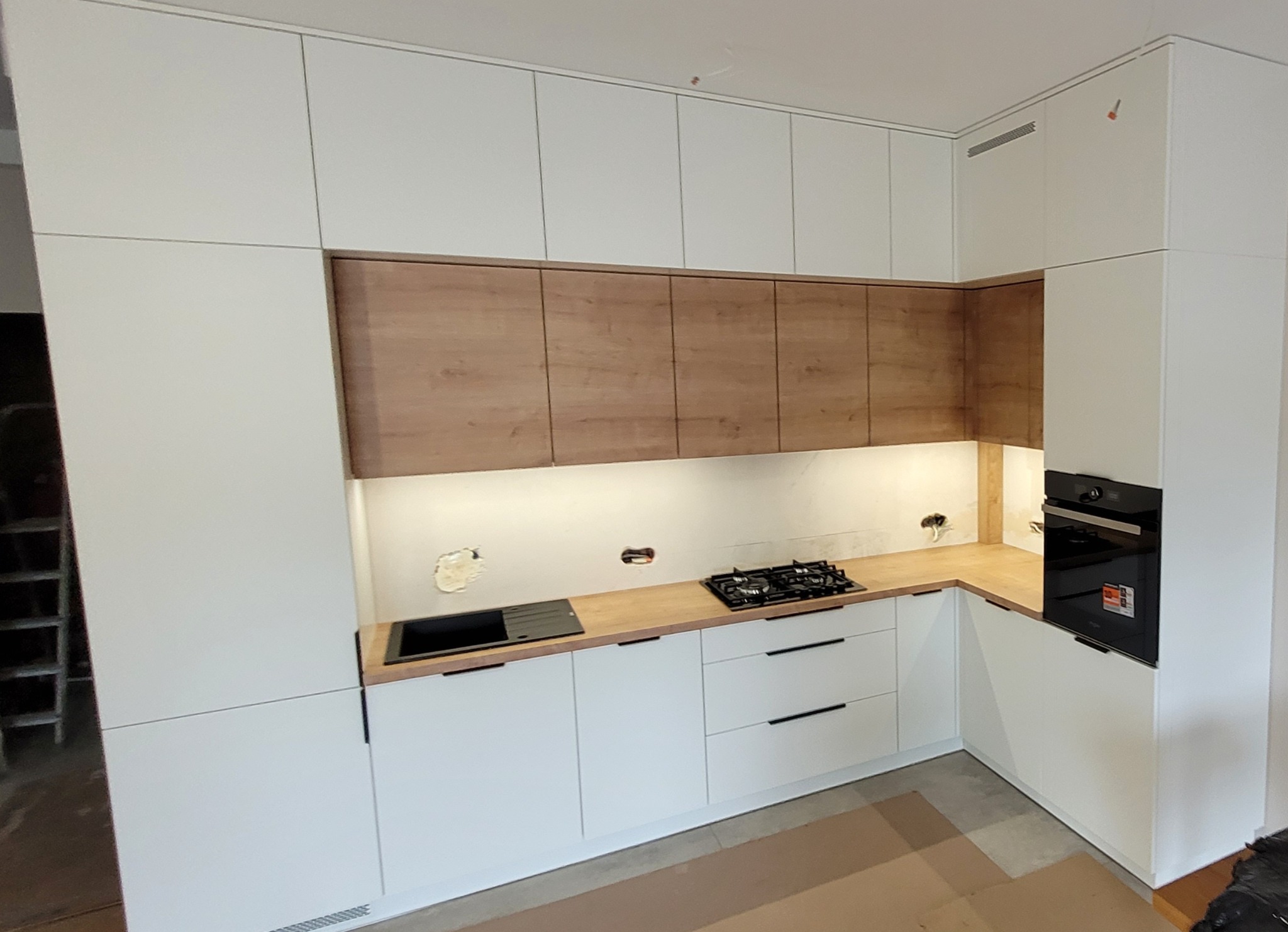 Nowoczesna kuchnia z białymi szafkami dolnymi i górnymi, drewnianymi szafkami wiszącymi, drewnianym blatem, czarną płytą grzewczą i piekarnikiem w zabudowie; widoczne niedokończone wykończenia ścian.
