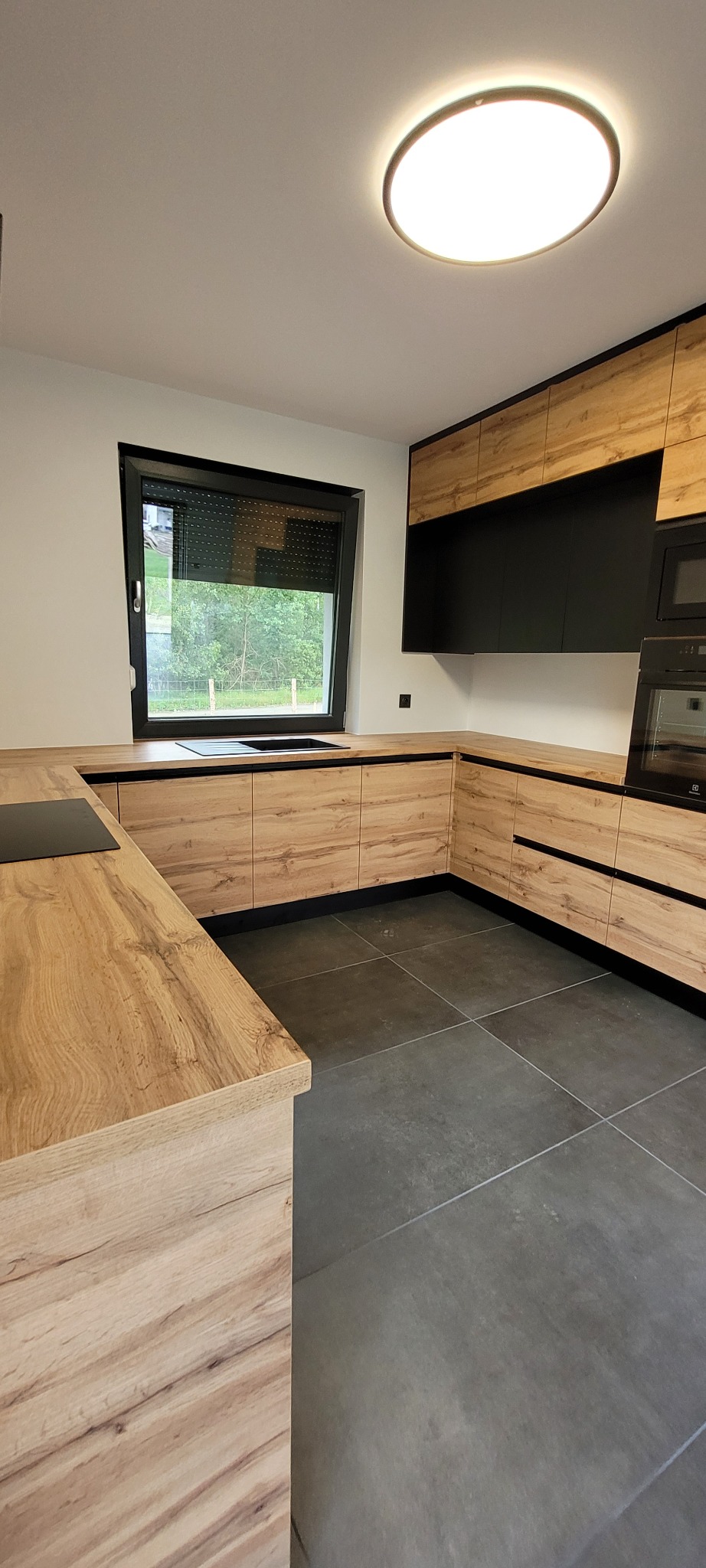 Nowoczesna kuchnia z drewnianymi frontami szafek i czarnymi akcentami, widok na okno z roletą i płytę indukcyjną na blacie.