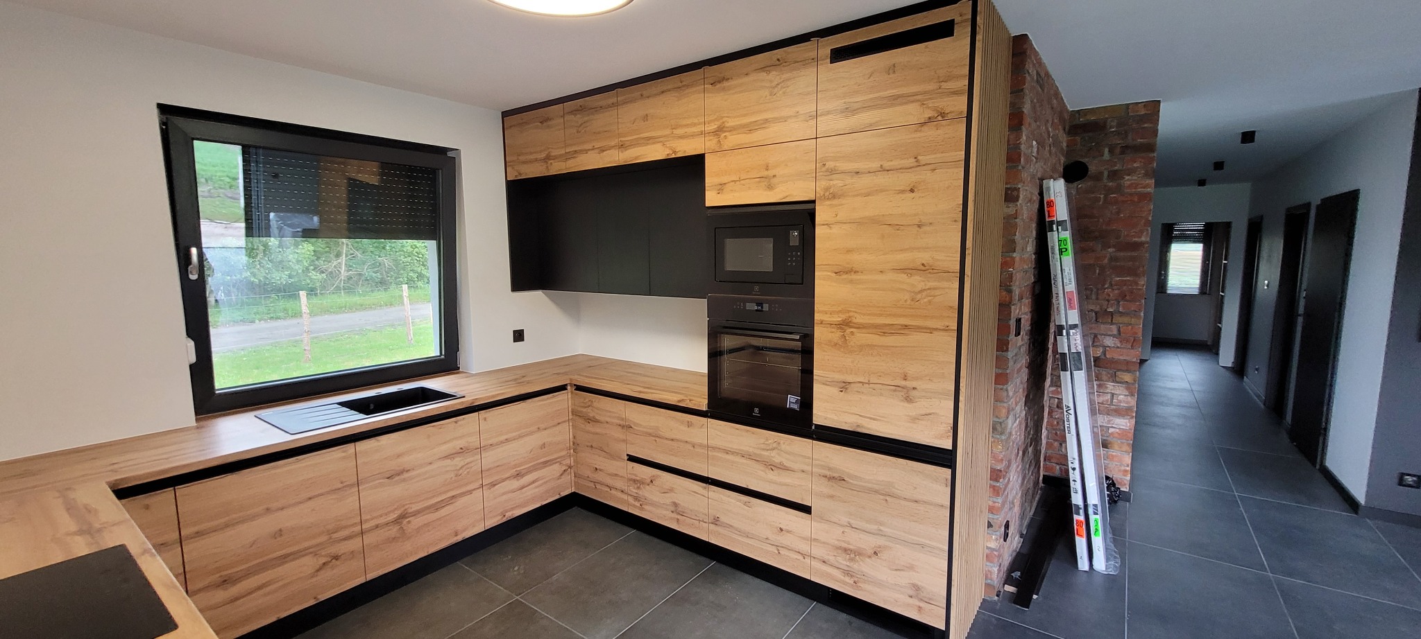 Nowoczesna kuchnia z drewnianymi frontami i czarnymi akcentami, w tle ceglana ściana i widok na zewnątrz przez okno.