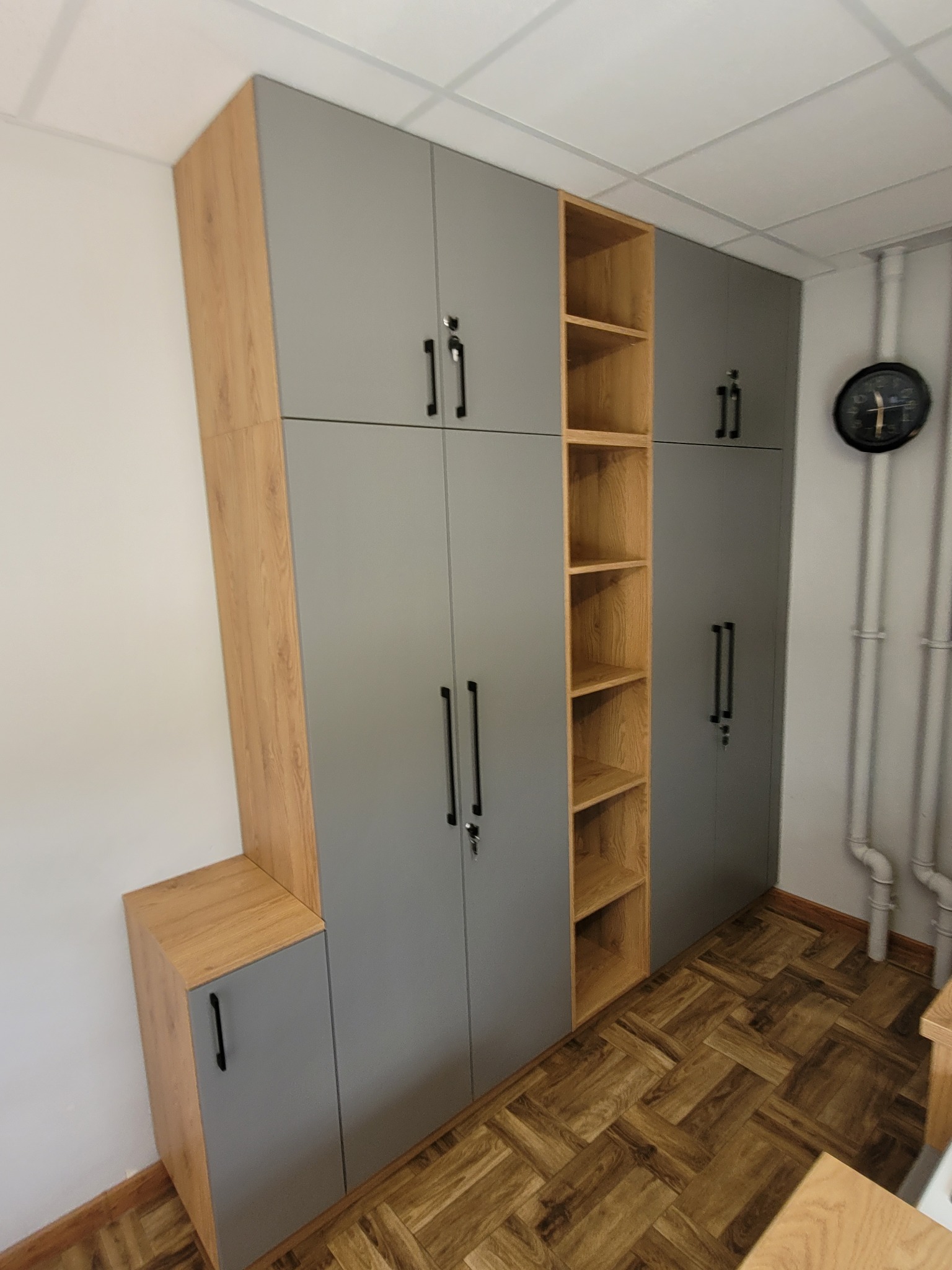 Szara szafa wnękowa z drewnianymi akcentami i otwartymi półkami, zamek w drzwiach, widok na parkiet i rury grzewcze.