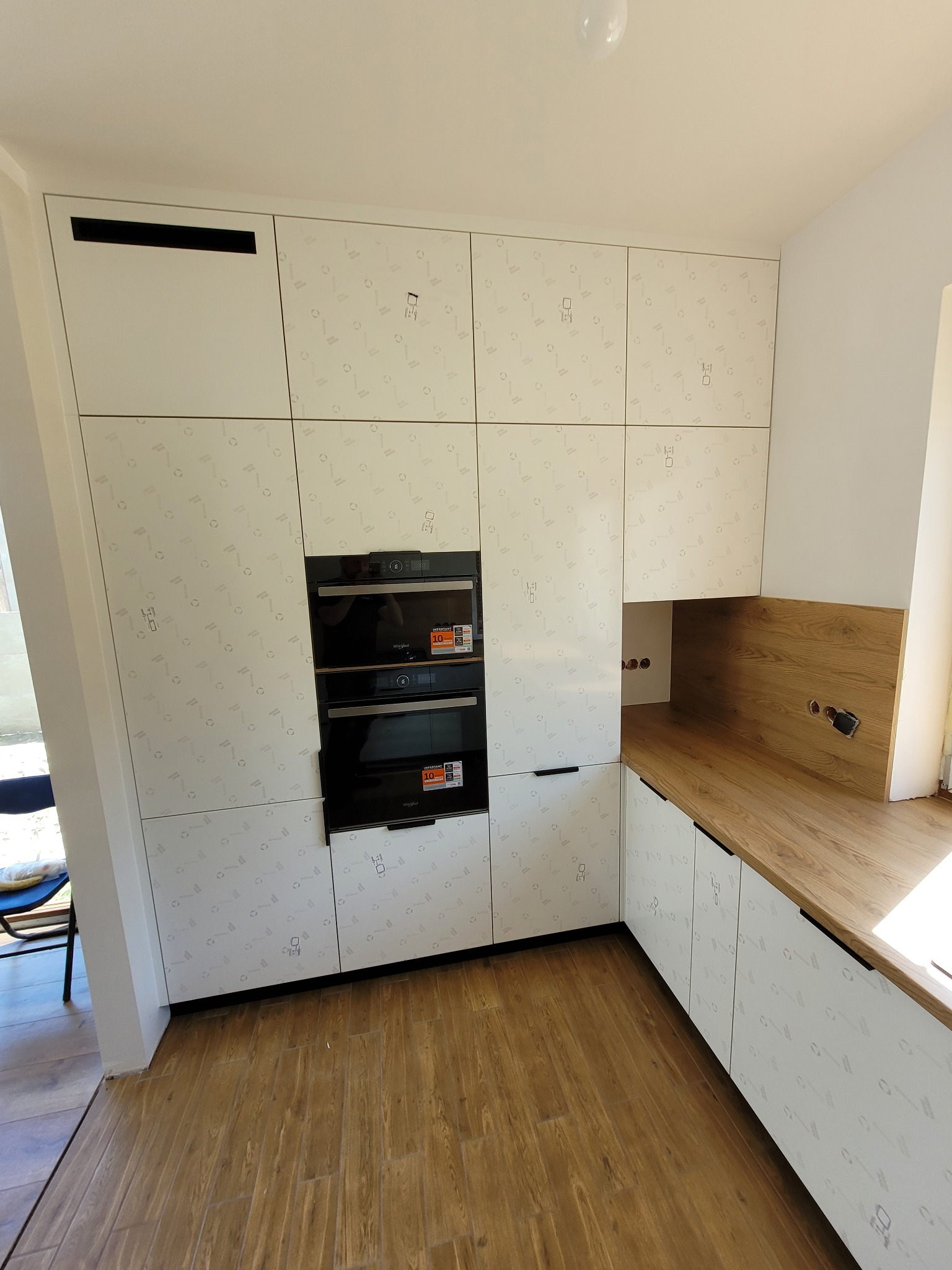 Nowoczesna kuchnia z białymi szafkami z delikatnym wzorem, wbudowanymi piekarnikami i drewnianym blatem, widok z perspektywy podłogi.