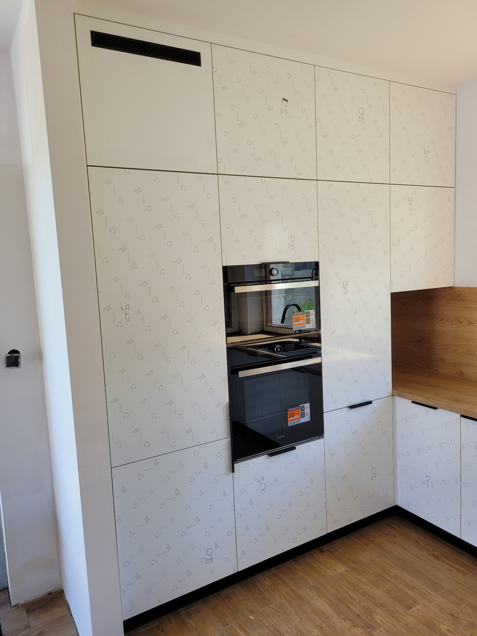 Biała zabudowa kuchenna z piekarnikiem i mikrofalówką, fronty z delikatnym wzorem, drewniany blat w rogu, widoczne ślady po remoncie na ścianie.