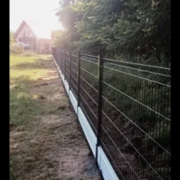 Długi, czarny panelowy system ogrodzeniowy z podmurówką w kolorze białym, biegnący wzdłuż trawnika, z domem w tle i drzewami w słoneczny dzień.