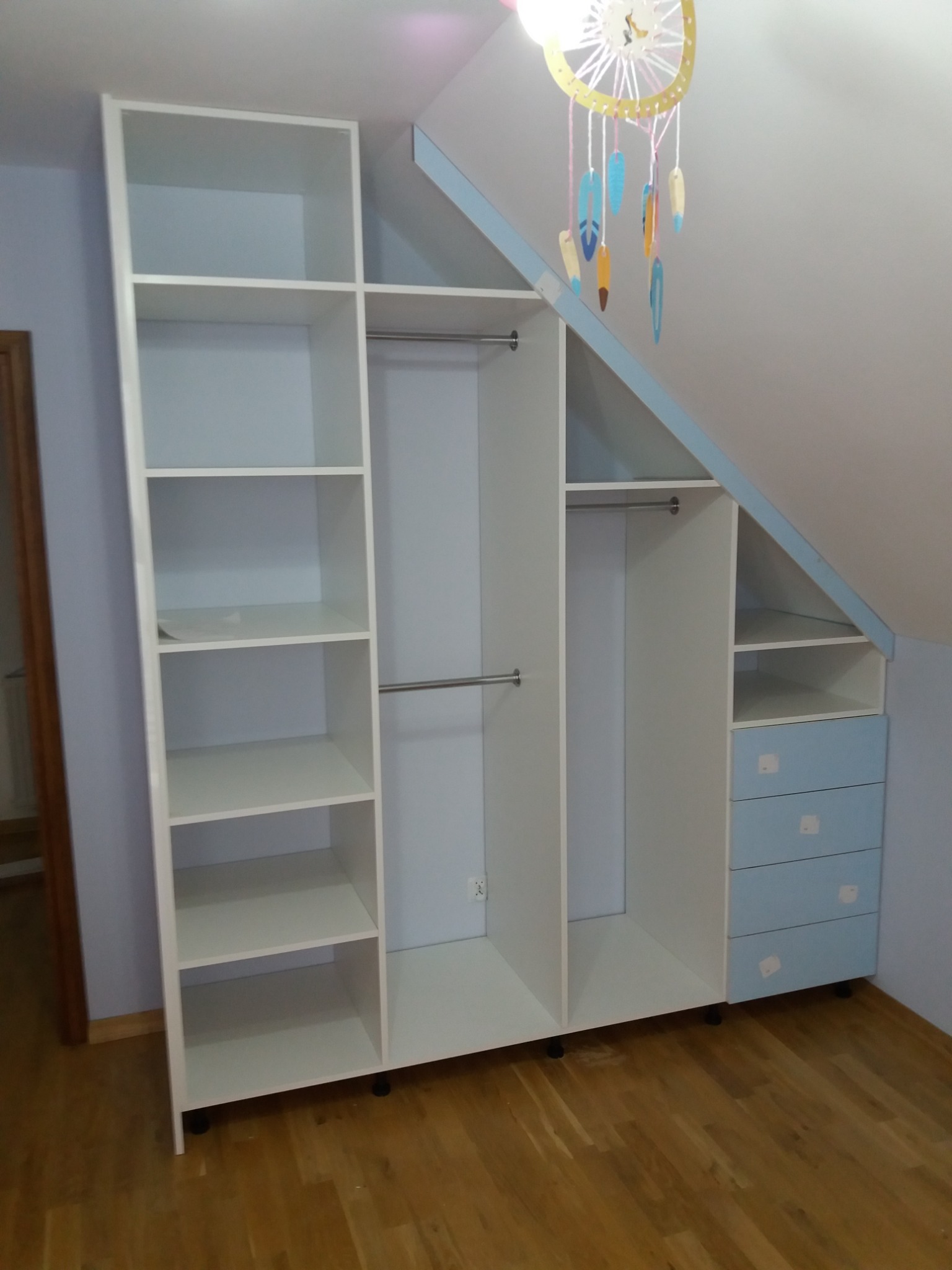 Biała szafa wnękowa z niebieskimi akcentami, dopasowana do skosu dachu, z półkami, drążkami na ubrania i szufladami, na drewnianej podłodze, z dekoracyjnym łapaczem snów w tle.