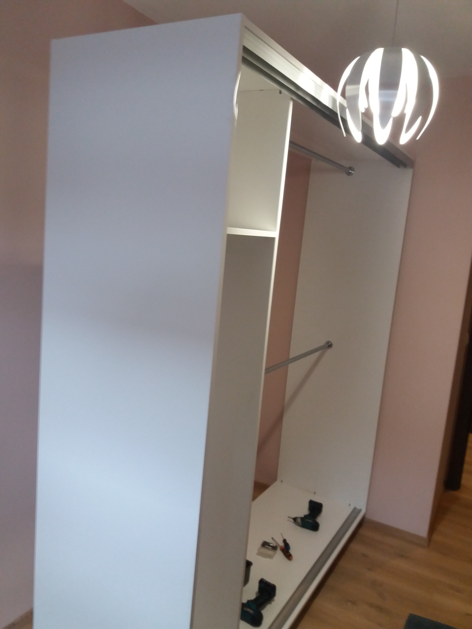 Montaż białej szafy przesuwnej z widocznymi narzędziami na podłodze i lampą w tle.