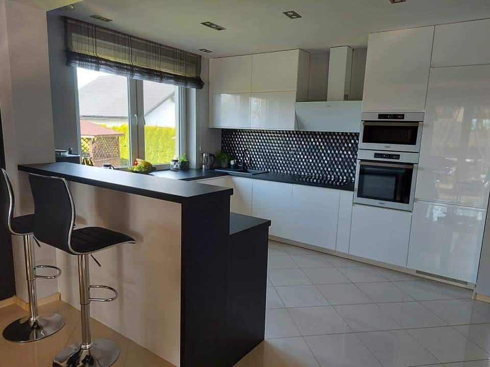 Nowoczesna, jasna kuchnia z białymi, lakierowanymi frontami, czarnym blatem, mozaiką w romby nad blatem i barkiem z dwoma hokerami.