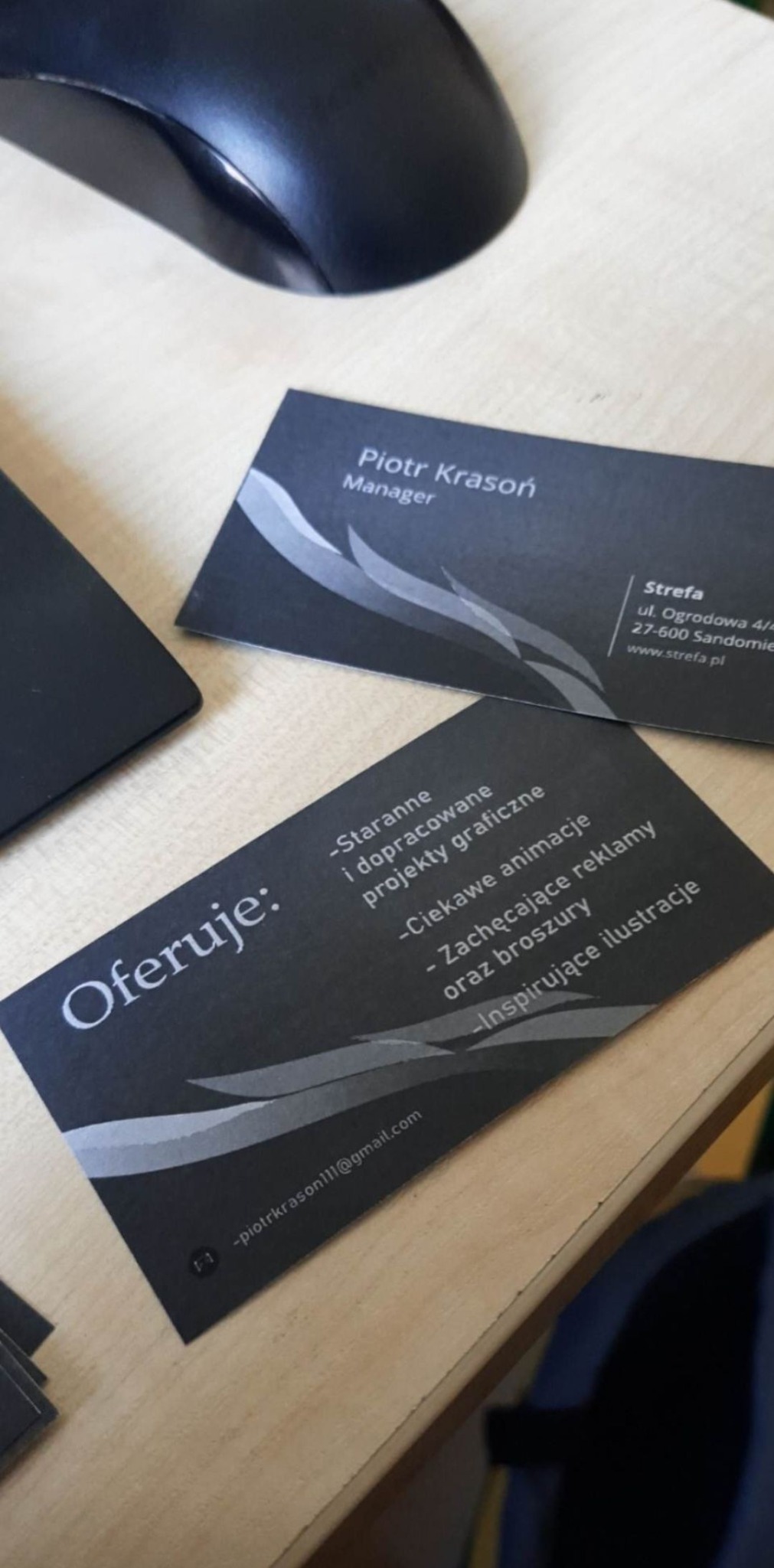 Stos wizytówek w ciemnych barwach na jasnym biurku, obok czarna myszka komputerowa. Wizytówki z danymi kontaktowymi i ofertą: staranne projekty graficzne, ciekawe animacje, zachęcające reklamy...