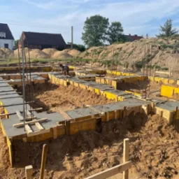 Wylane betonowe ławy fundamentowe w żółtym szalunku, widoczne wystające pręty zbrojeniowe i pracownik przy dalszych pracach, w tle usypana ziemia i zabudowa jednorodzinna.