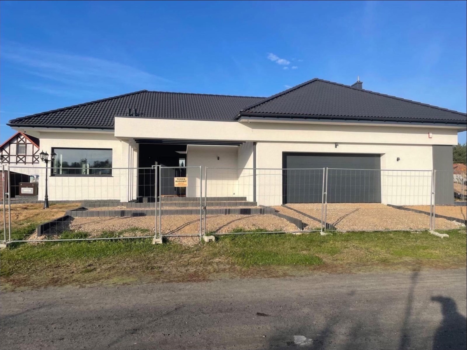 Nowoczesny dom jednorodzinny z garażem w trakcie wykończenia, otoczony tymczasowym ogrodzeniem budowlanym i kamienistym podłożem, widoczny znak 'Teren Budowy'.