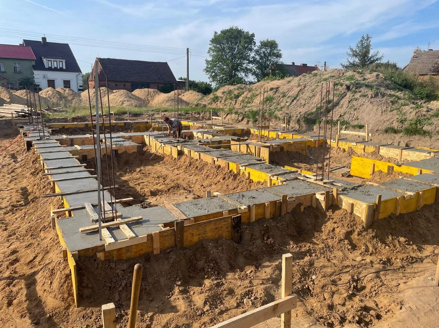 Wylane betonowe ławy fundamentowe w żółtym szalunku, widoczne wystające pręty zbrojeniowe i pracownik przy dalszych pracach, w tle usypana ziemia i zabudowa jednorodzinna.