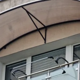 Zadaszenie balkonowe z poliwęglanu, wsparte na metalowej konstrukcji, widoczne z zewnątrz budynku.