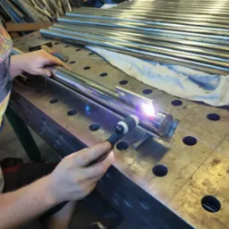 Praca przy użyciu spawarki TIG na metalowym profilu, widoczny spawacz w masce ochronnej z motywem płomieni, warsztatowa przestrzeń robocza z ułożonymi rurami w tle.