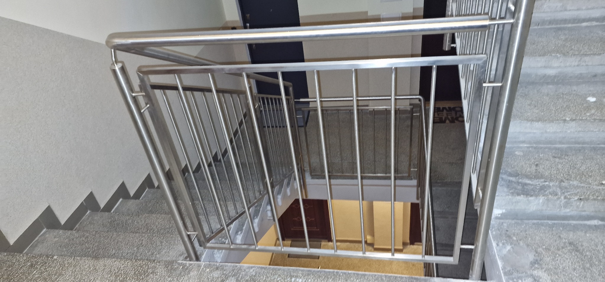 Klatka schodowa z szarymi, betonowymi schodami i metalową, nierdzewną balustradą z pionowymi prętami, widok z góry.