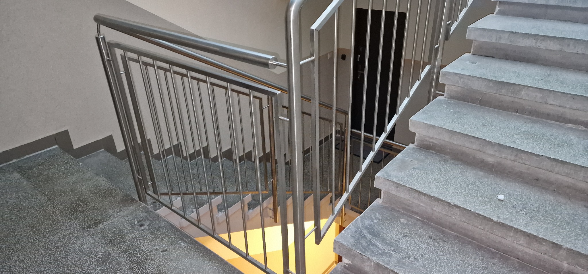 Wewnętrzne schody z szarego kamienia z metalową balustradą z pionowymi prętami, widok z góry na dół, z widocznym fragmentem podłogi poniżej i czarnymi drzwiami w tle.
