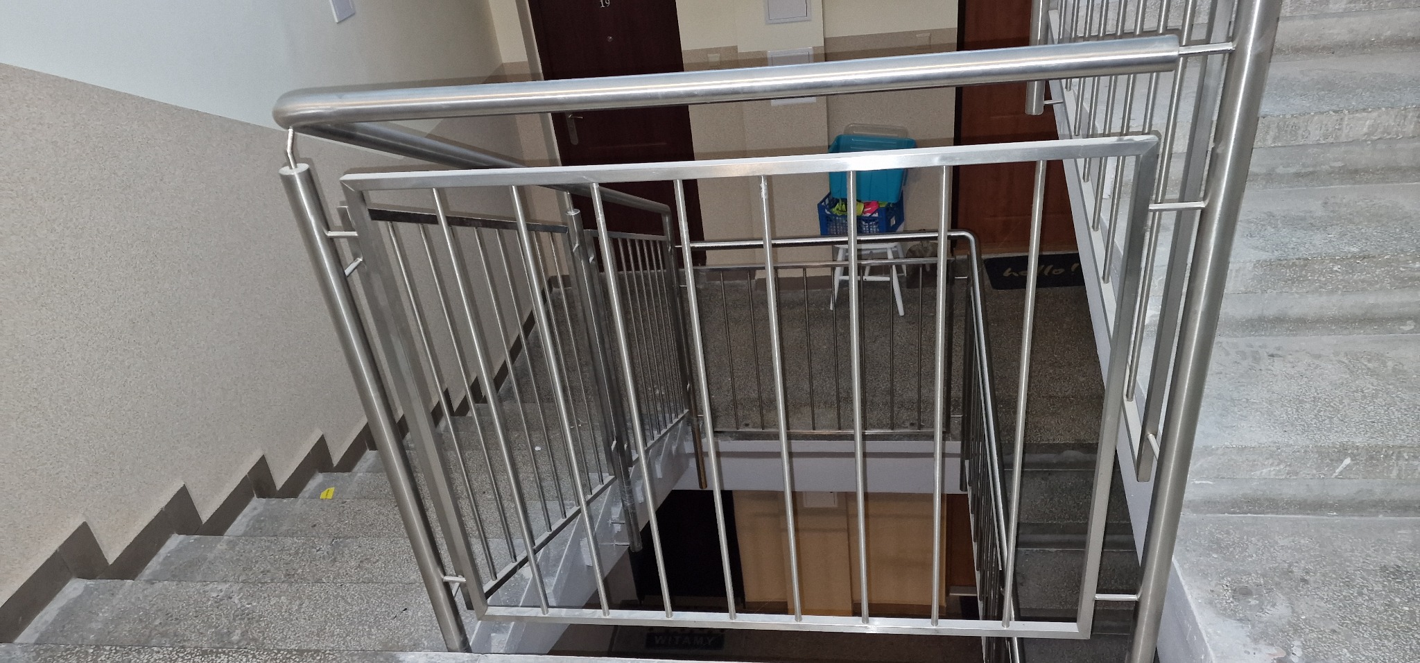 Nierdzewna balustrada schodowa z pionowymi prętami, widok z góry na schody w bloku mieszkalnym, z widocznymi drzwiami do mieszkań na każdym piętrze.