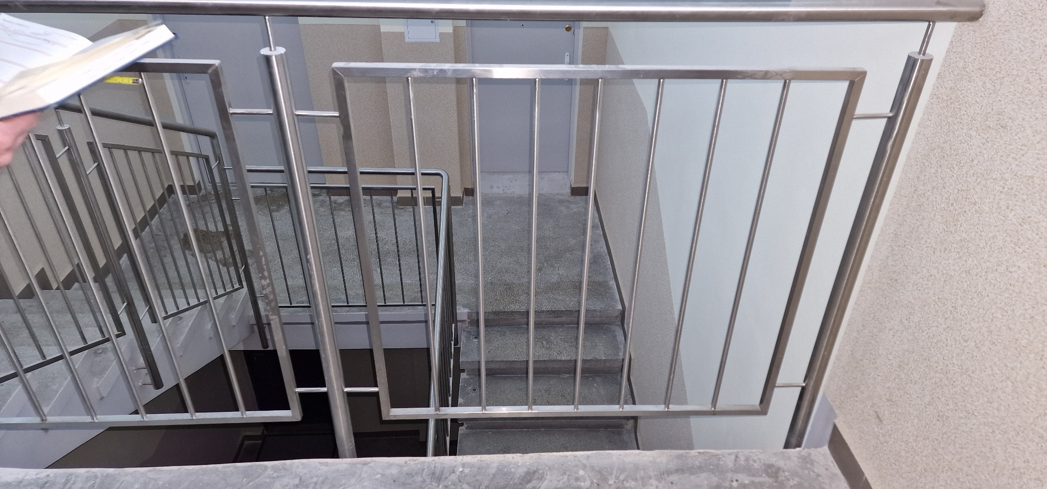 Wewnętrzne schody z poręczami ze stali nierdzewnej, widok z góry na kilka stopni, z fragmentem otwartej książki w lewym górnym rogu.