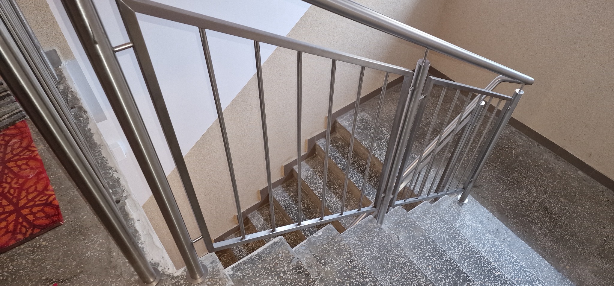 Widok z góry na schody z szarymi, lastrykowymi stopniami i metalową poręczą ze stali nierdzewnej, zamontowaną w klatce schodowej o ścianach w kolorze złamanej bieli i beżu.