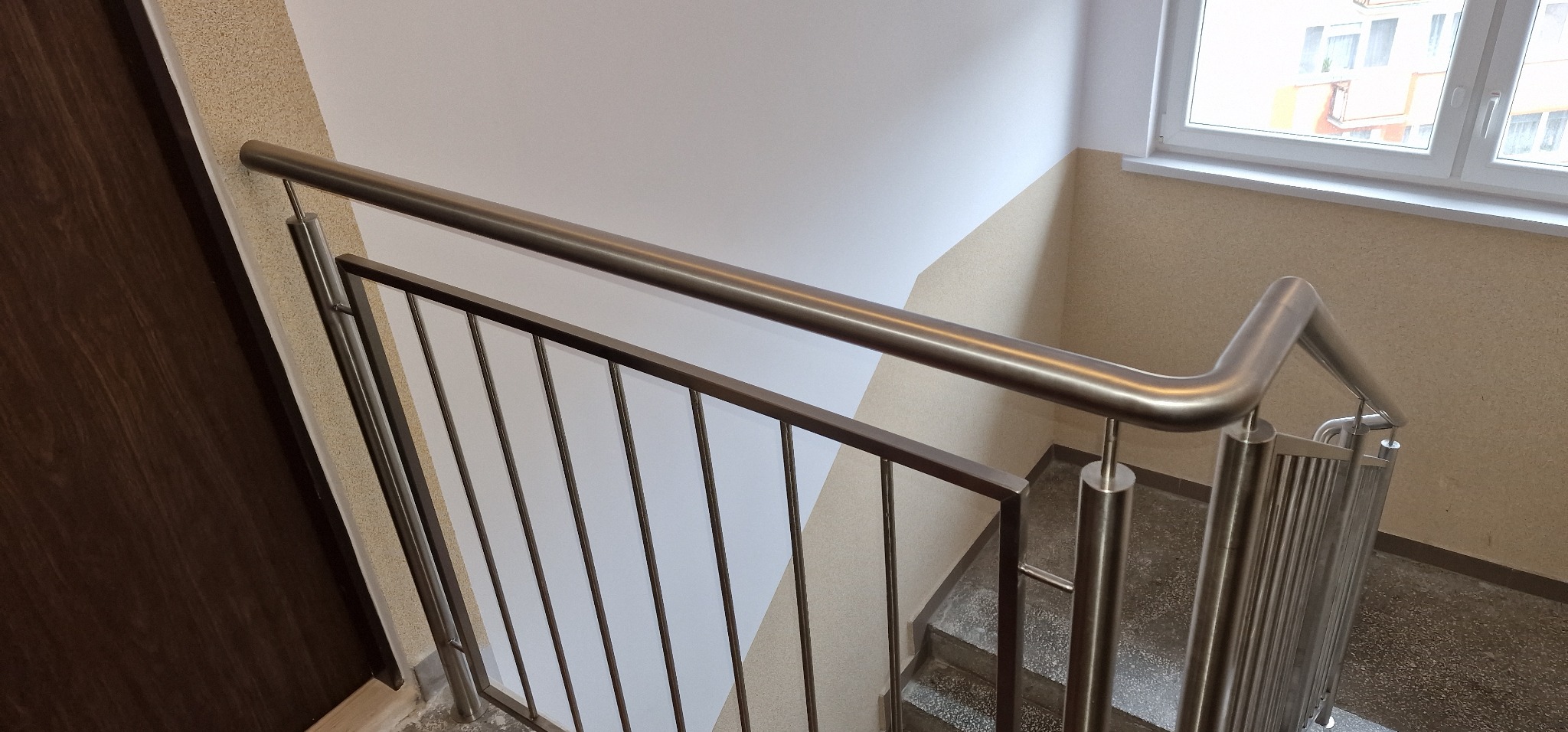 Nierdzewna poręcz schodowa z pionowymi wypełnieniami, widok z góry na schody z szarymi stopniami i beżową ścianą.