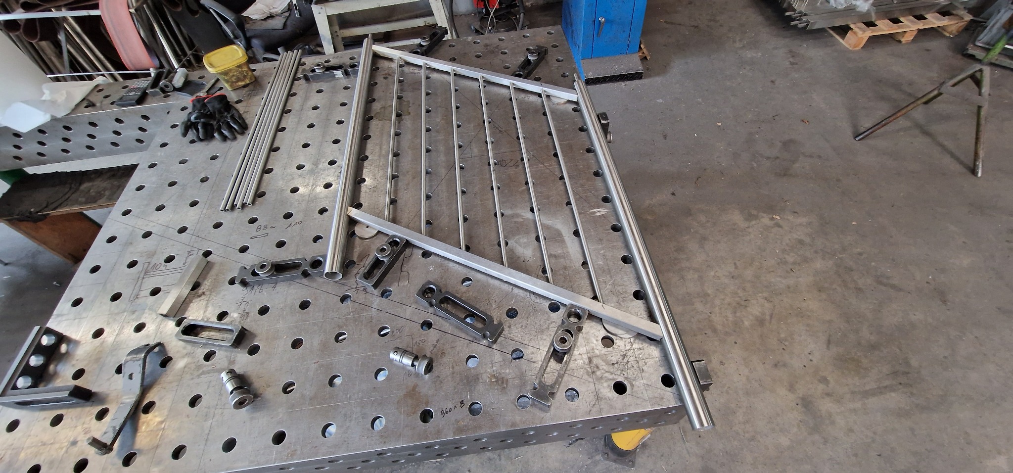 Fragment balustrady nierdzewnej w trakcie spawania na stole spawalniczym z widocznymi narzędziami i elementami mocującymi.