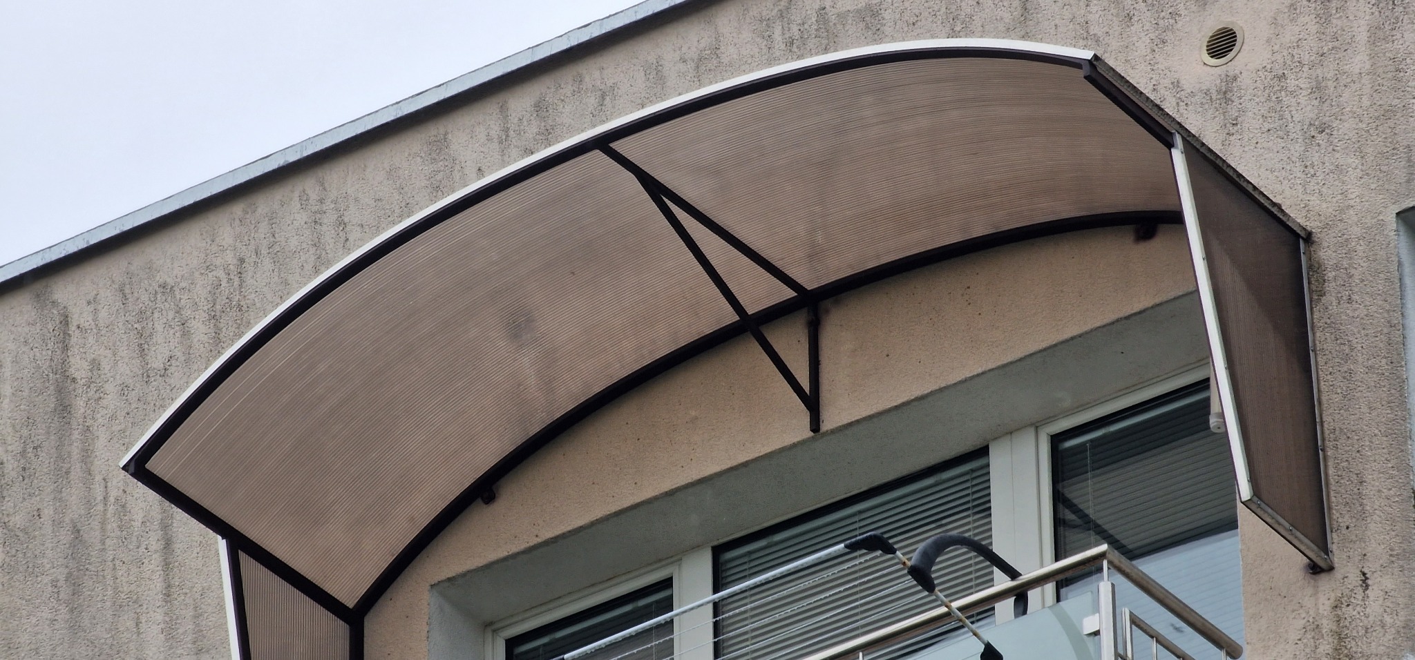 Zadaszenie nad balkonem z poliwęglanu, wsparte na metalowej konstrukcji, widoczne z dołu, z fragmentem balustrady balkonowej w kadrze.