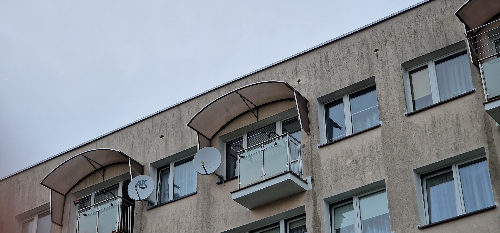 Szare balkony z balustradami ze stali nierdzewnej i szkła, zadaszone daszkami z poliwęglanu, na tle budynku z elewacją w odcieniach szarości i antenami satelitarnymi.