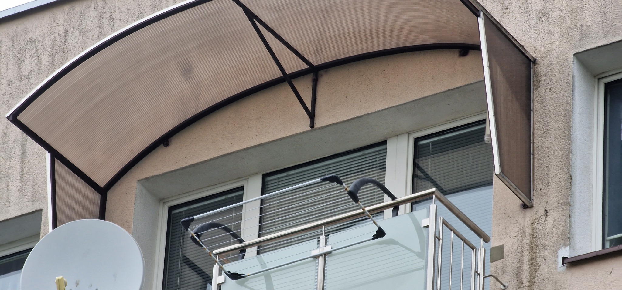 Zadaszenie balkonu z poliwęglanu na metalowej konstrukcji, widoczny balkon z szklaną balustradą i suszarką na pranie, antena satelitarna na ścianie budynku.
