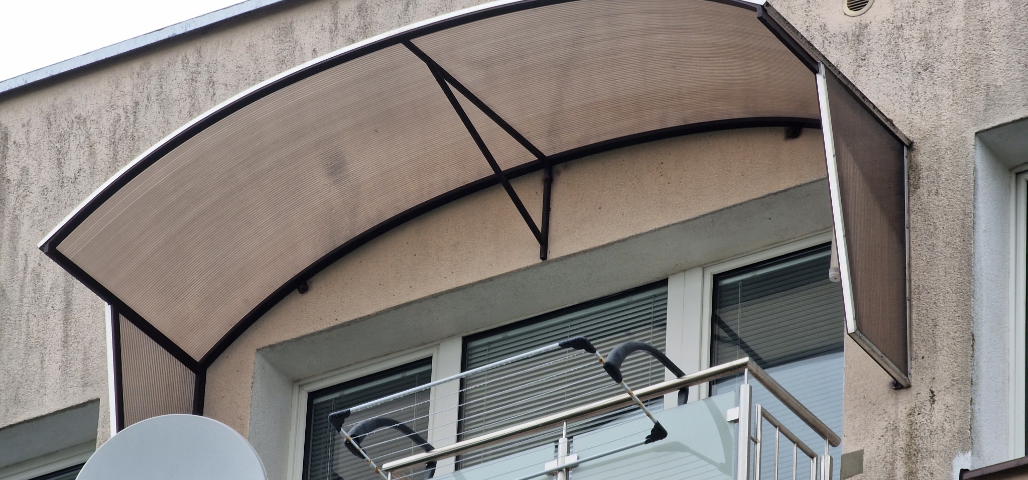 Zadaszenie balkonowe z poliwęglanu, wsparte na metalowej konstrukcji, widoczne z zewnątrz budynku.