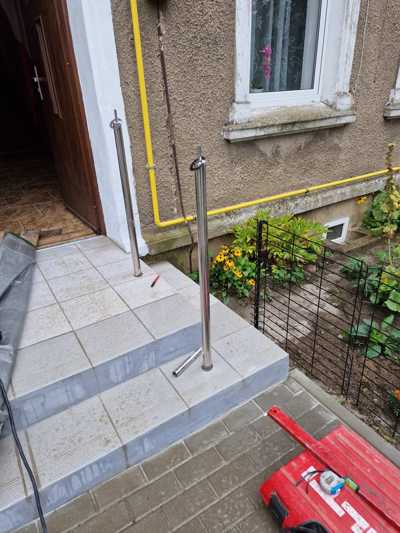 Montaż poręczy nierdzewnych na schodach wejściowych do domu, widoczne słupki przed przytwierdzeniem, narzędzia pracy obok.
