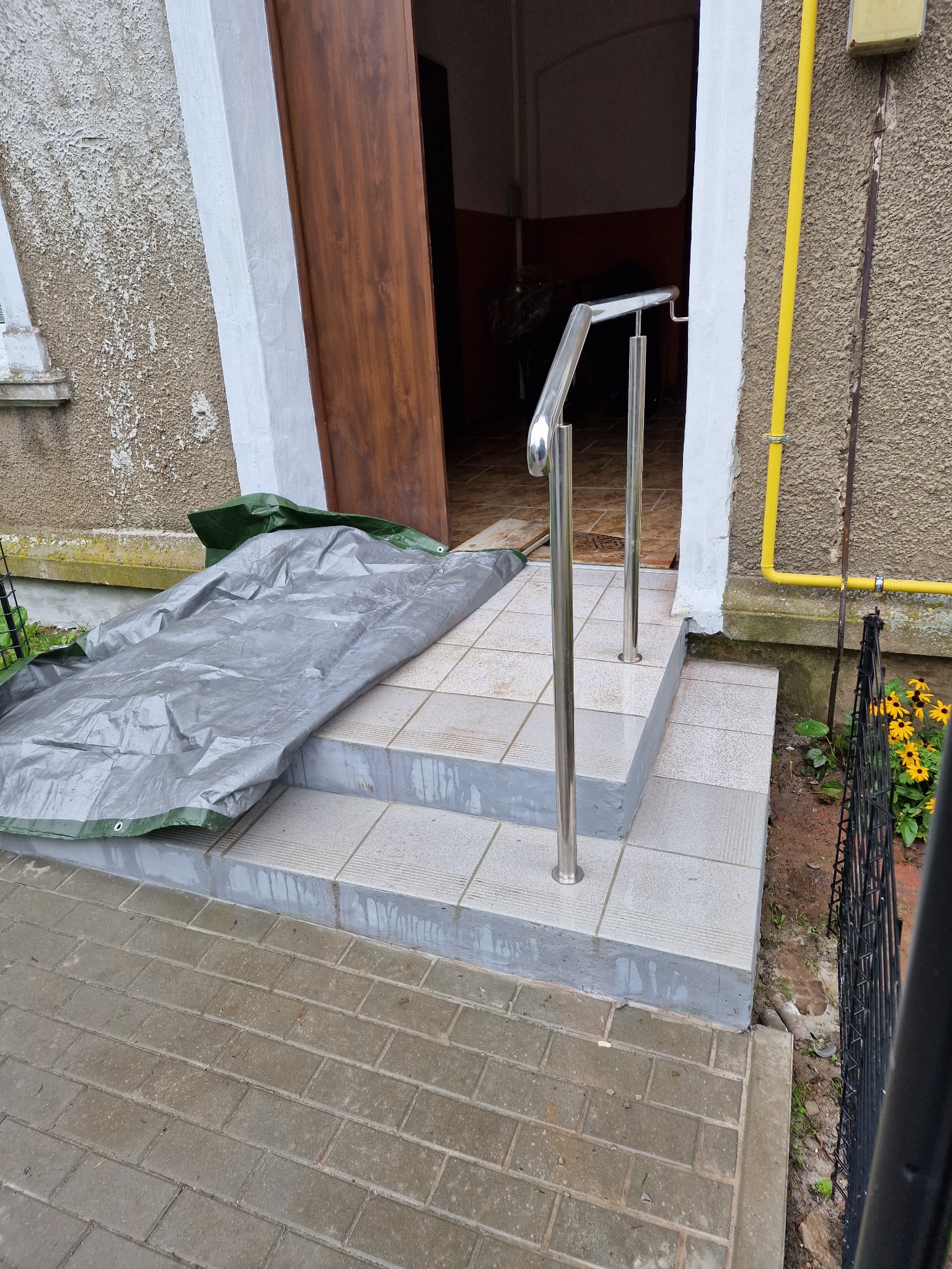 Zewnętrzne schody wejściowe z trzema stopniami wyłożone płytkami, z zamontowaną prostą, metalową poręczą po prawej stronie, częściowo zakryte szarą plandeką, na tle elewacji budynku z żółtą rurą...