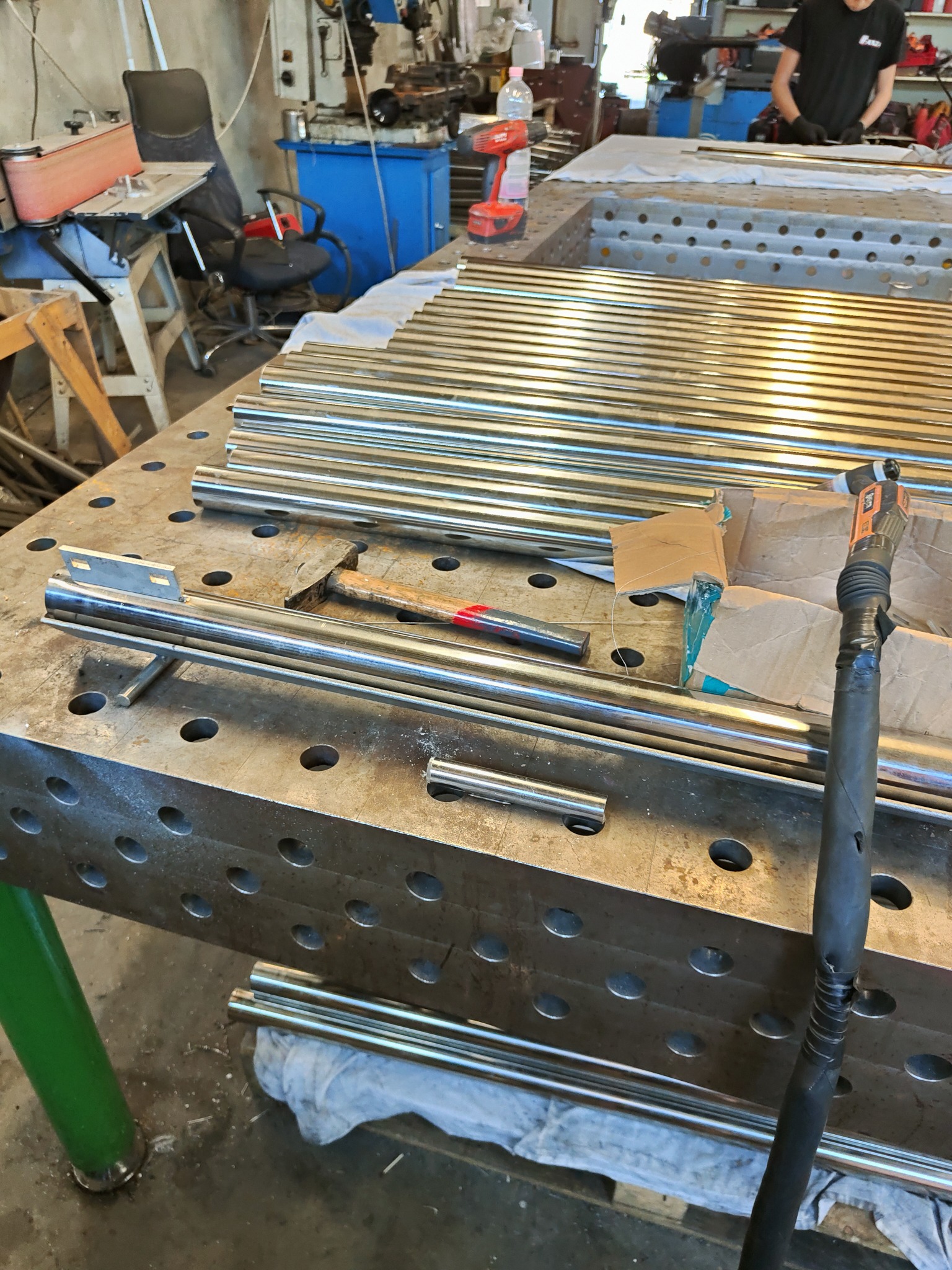 Przygotowane do obróbki rury ze stali nierdzewnej leżące na perforowanym stole warsztatowym, obok młotek i narzędzie pneumatyczne, widoczny fragment pracownika w tle.