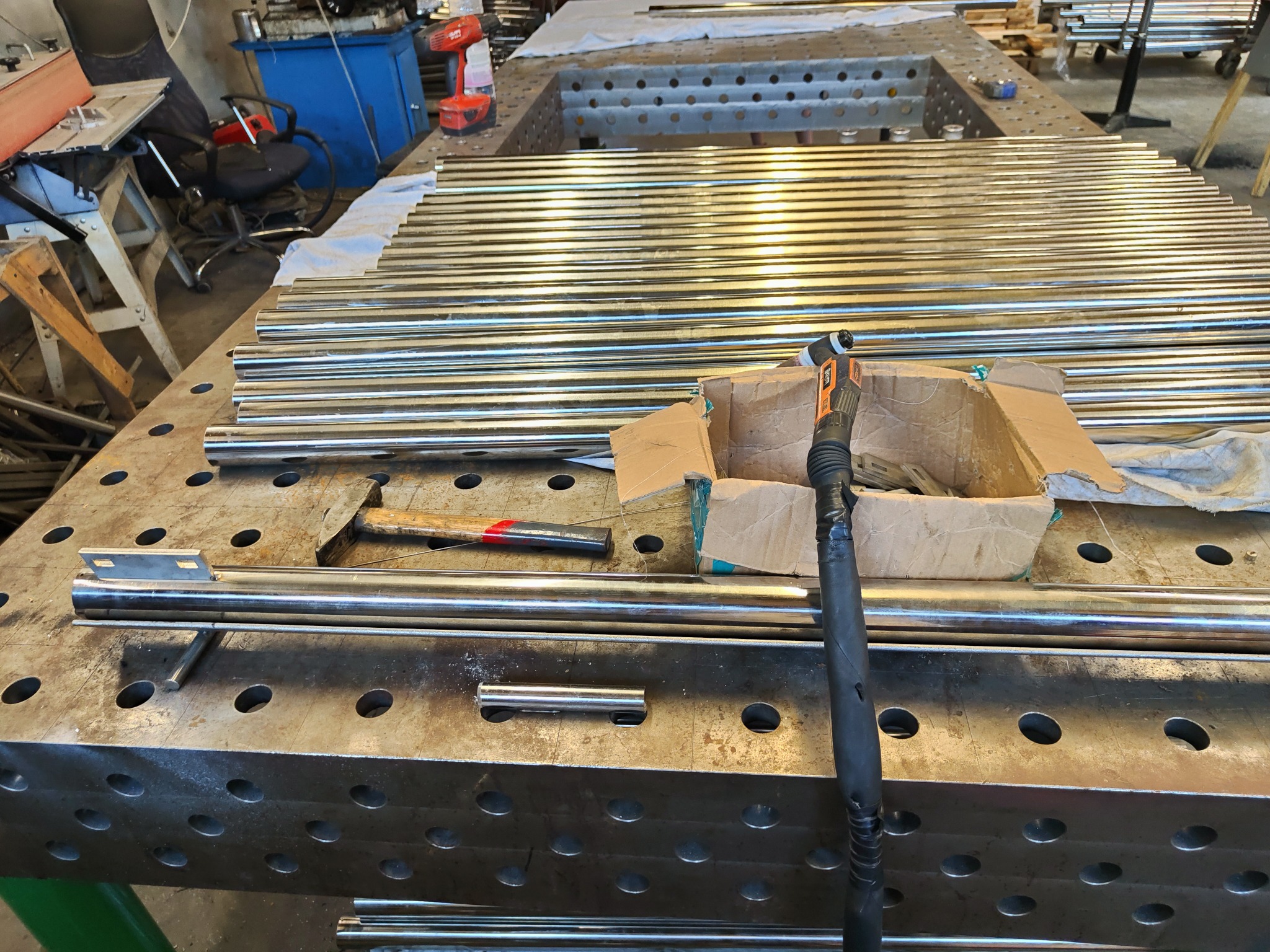 Przygotowane do obróbki rury ze stali nierdzewnej leżące na perforowanym stole warsztatowym, obok młotek i uchwyt do spawarki w kartonowym pudełku.