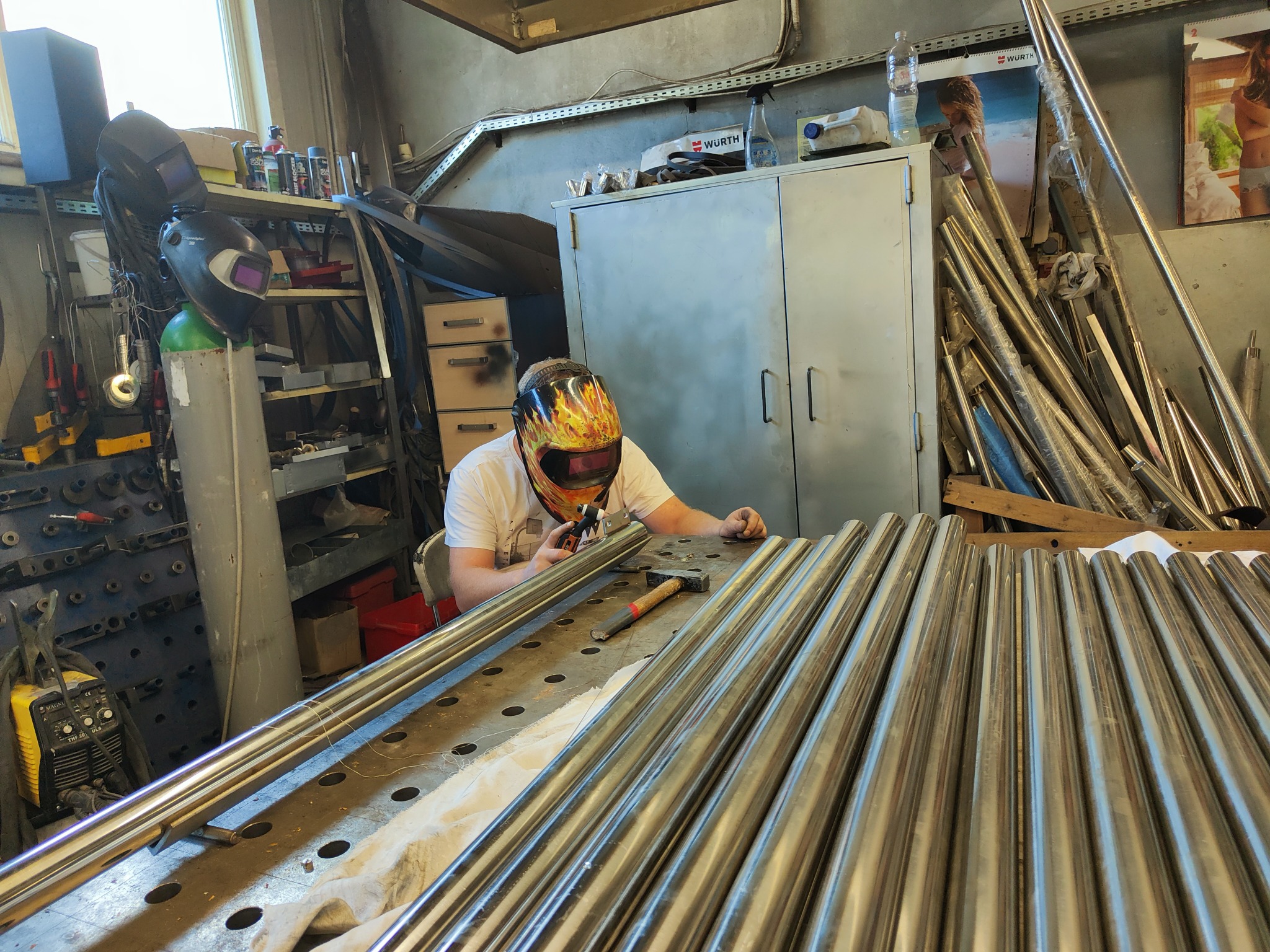 Pracownik w masce spawalniczej z motywem płomieni przygotowuje elementy z rur nierdzewnych na perforowanym stole warsztatowym, w tle widoczny sprzęt i akcesoria warsztatowe oraz metalowe profile.