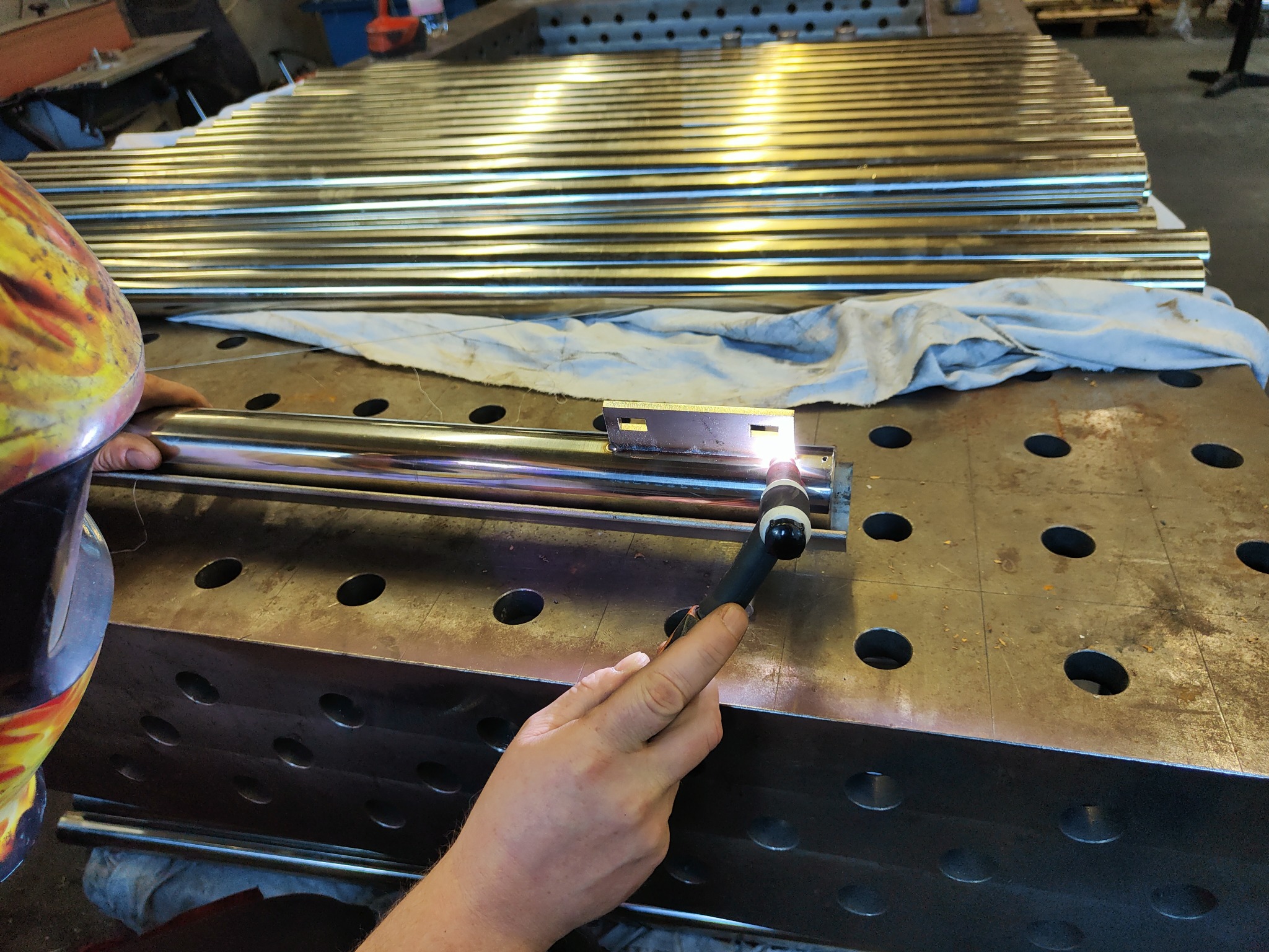 Praca przy łączeniu metalowych rur za pomocą spawarki TIG na perforowanym stole spawalniczym; w tle ułożone równolegle przygotowane elementy konstrukcyjne.