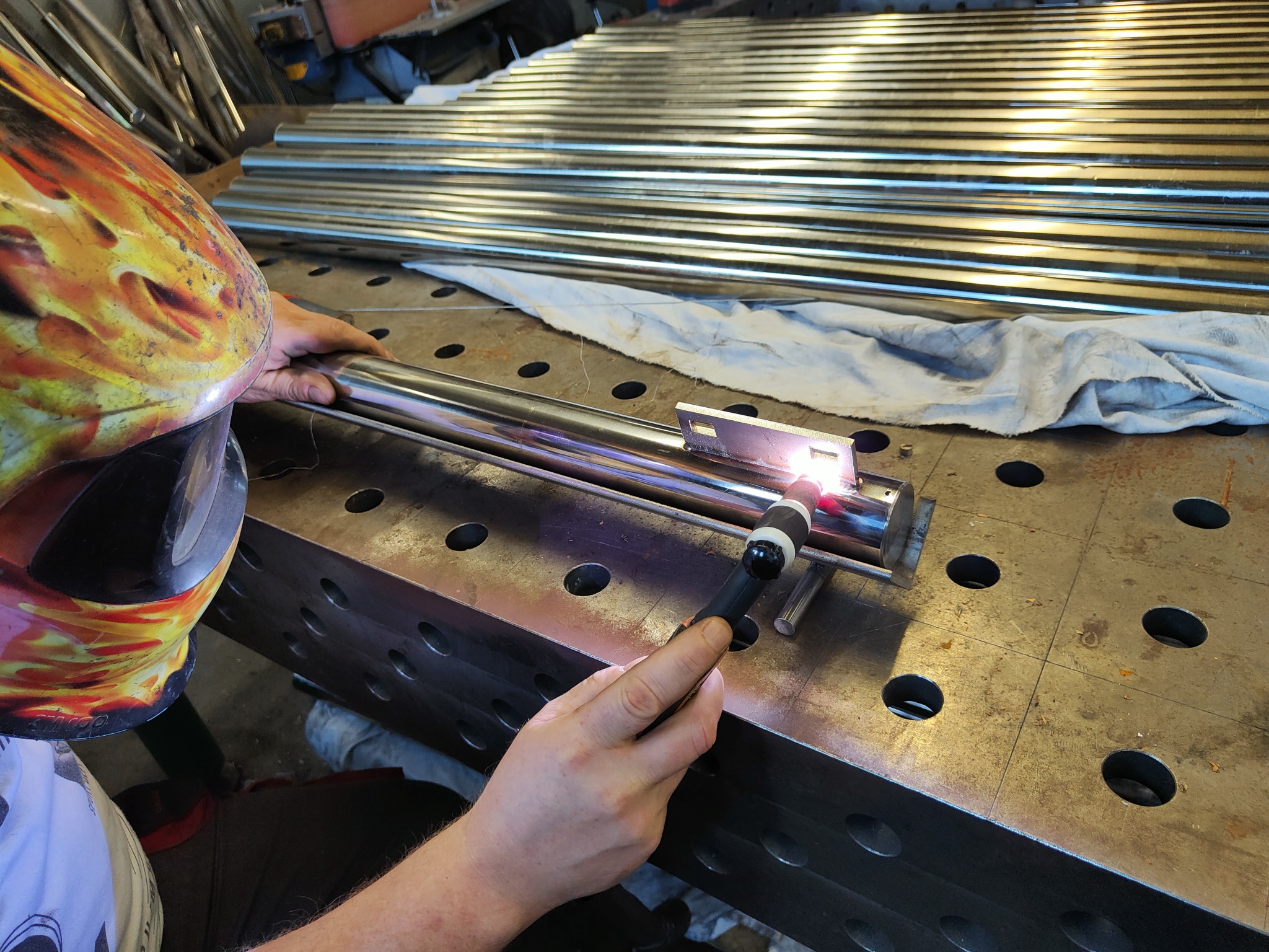 Praca przy spawaniu rury ze stali nierdzewnej na perforowanym stole spawalniczym, widoczna maska spawalnicza z motywem płomieni i rząd rur w tle.