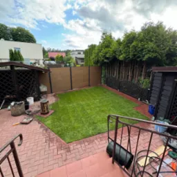 Widok z góry na świeżo położony trawnik z rolki w ogrodzie z drewnianym ogrodzeniem, brązową altaną i drzewami iglastymi w tle, widoczne narzędzia ogrodnicze na schodach i kostce brukowej.