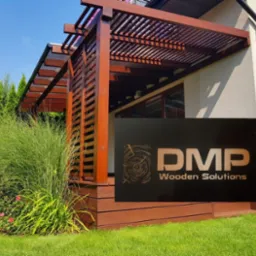 Drewniana pergola przylegająca do domu z widocznym logo firmy DMP Wooden Solutions na pierwszym planie, otoczona zielenią.