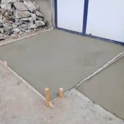 Świeżo wylana, szara posadzka w drewnianym szalunku, widoczny stos gruzu w tle i brama garażowa.