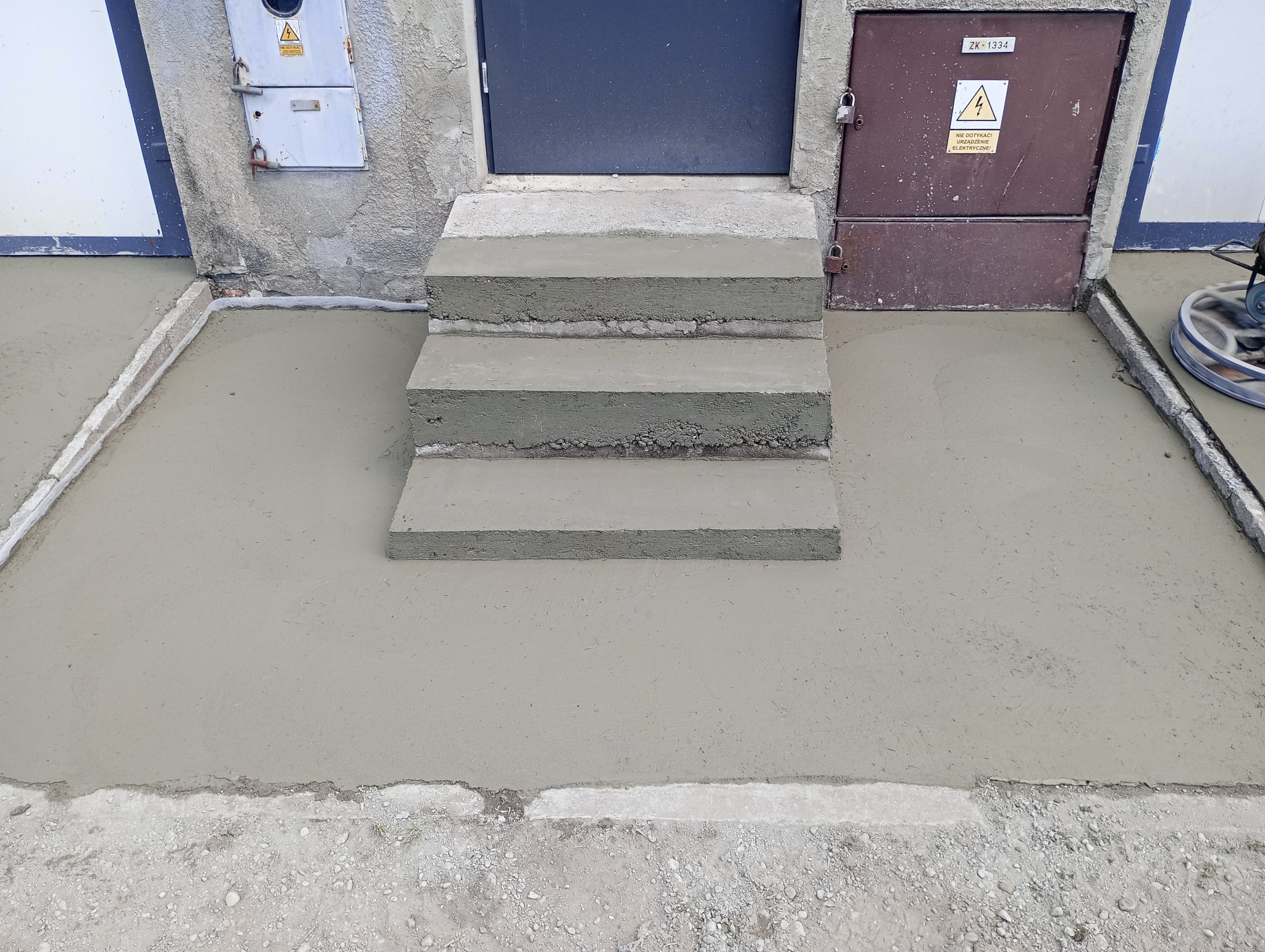 Świeżo wylana, gładka posadzka z jastrychu cementowego przed wejściem do budynku, z trzema betonowymi stopniami i widoczną maszyną do zacierania na krawędzi.