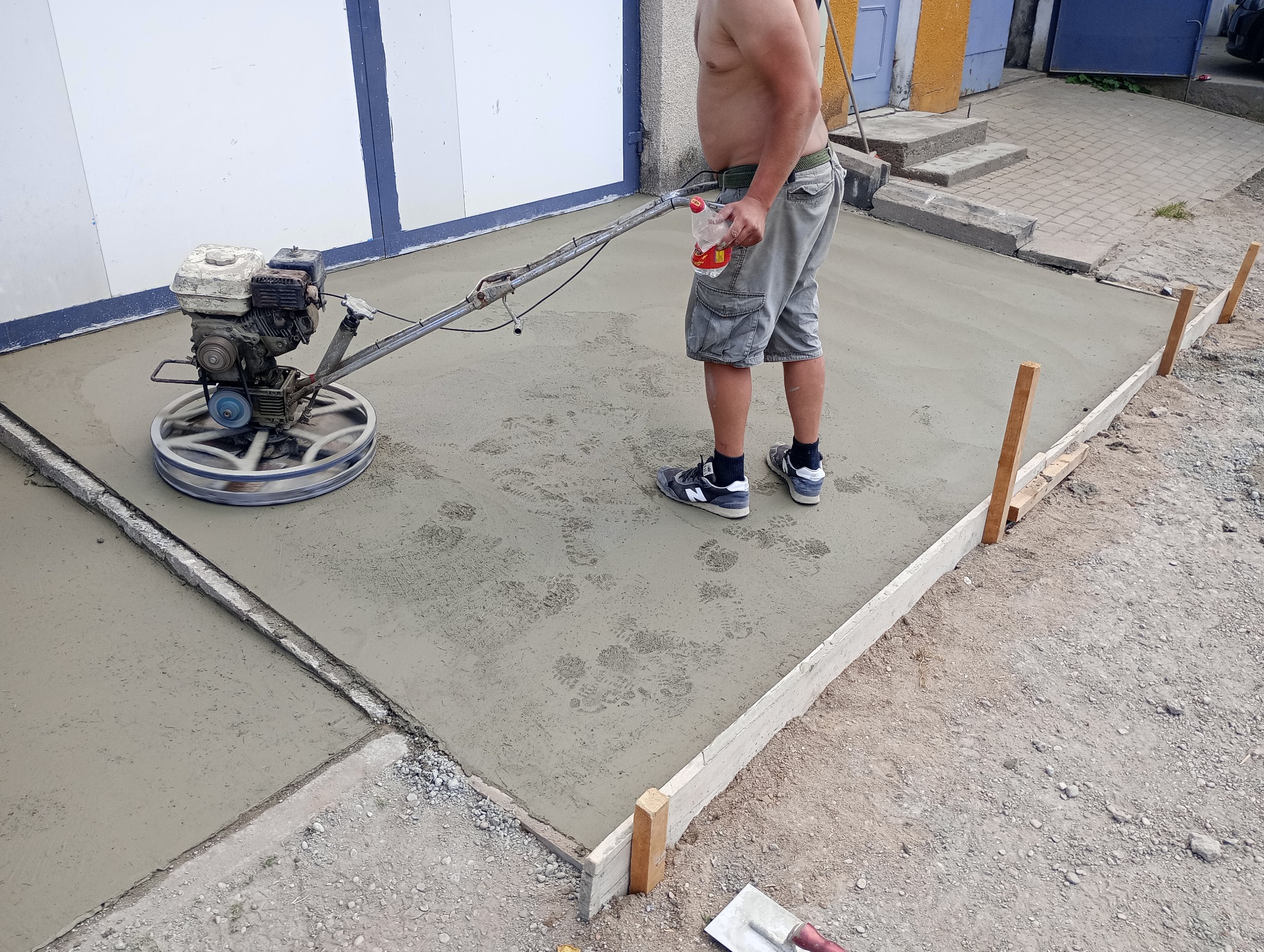 Wykończenie posadzki jastrychowej zacieraczką mechaniczną, widoczne ślady butów na świeżym betonie, drewniane szalunki i osoba z butelką w ręku.