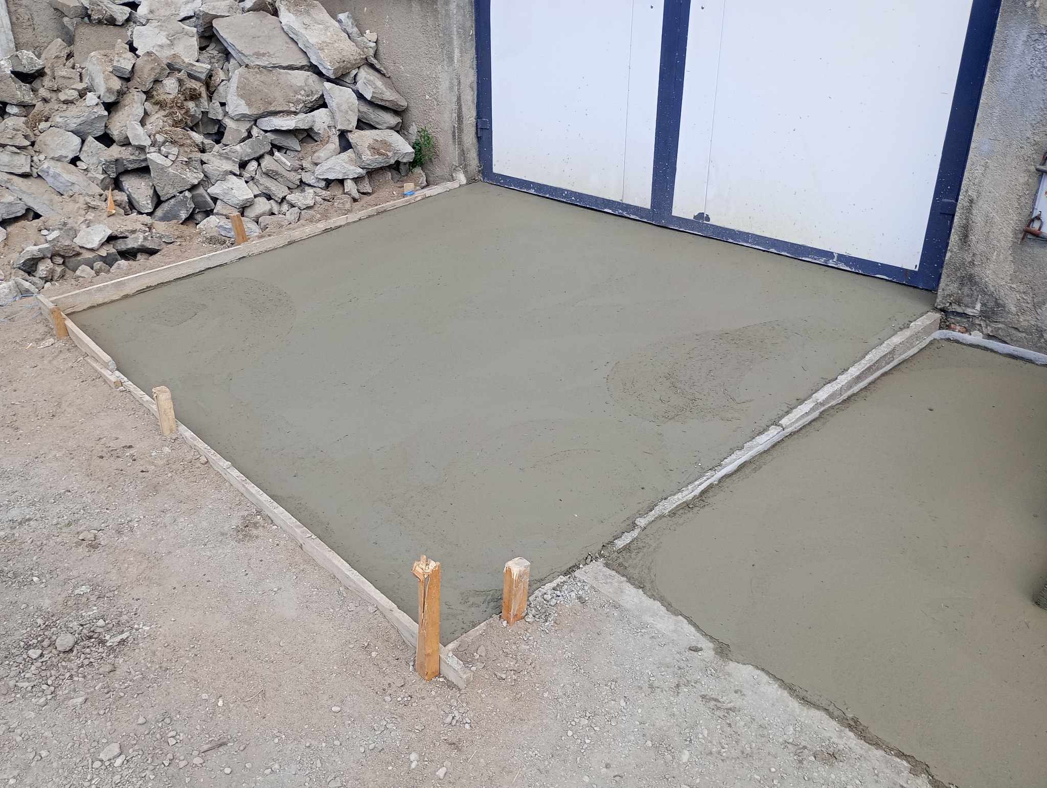 Świeżo wylana, szara posadzka w drewnianym szalunku, widoczny stos gruzu w tle i brama garażowa.