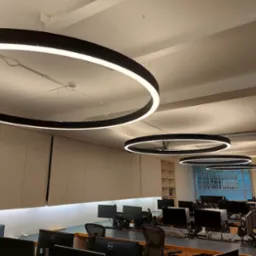 Biuro z rzędem okrągłych lamp LED o czarnej obudowie, zawieszonych pod sufitem, oświetlających stanowiska komputerowe.