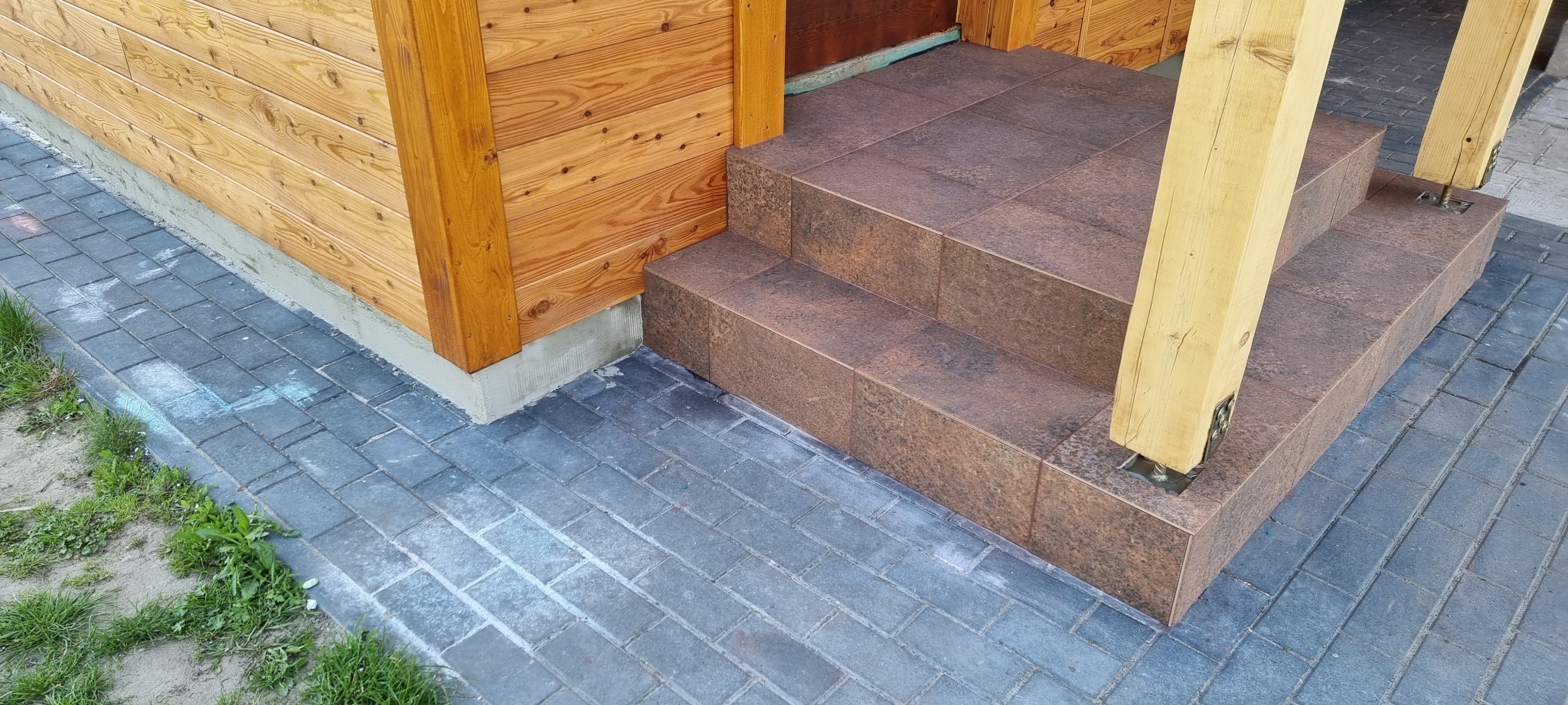 Schody wejściowe wyłożone brązowymi płytkami imitującymi kamień, z drewnianymi słupami podtrzymującymi zadaszenie, na tle drewnianej elewacji i szarej kostki brukowej.