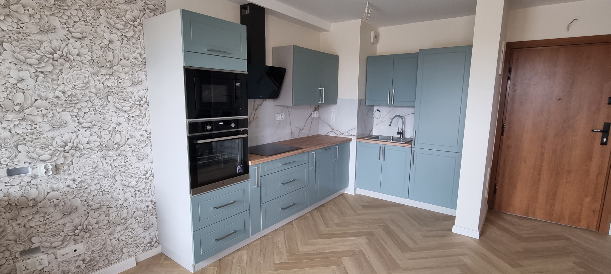 Zakończona aranżacja kuchni w odcieniach błękitu i drewna, z wzorzystą tapetą w kwiaty na jednej ze ścian i parkietem ułożonym w jodełkę.