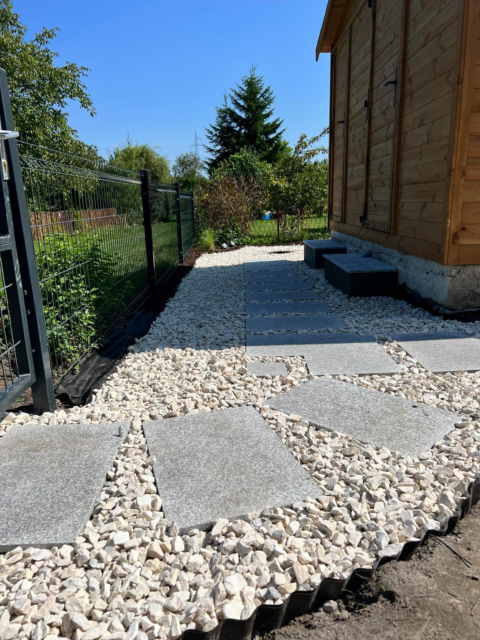 Ścieżka z szarych płyt ułożona na białym żwirze prowadząca do drewnianego domku, widoczne ogrodzenie i zieleń w tle.
