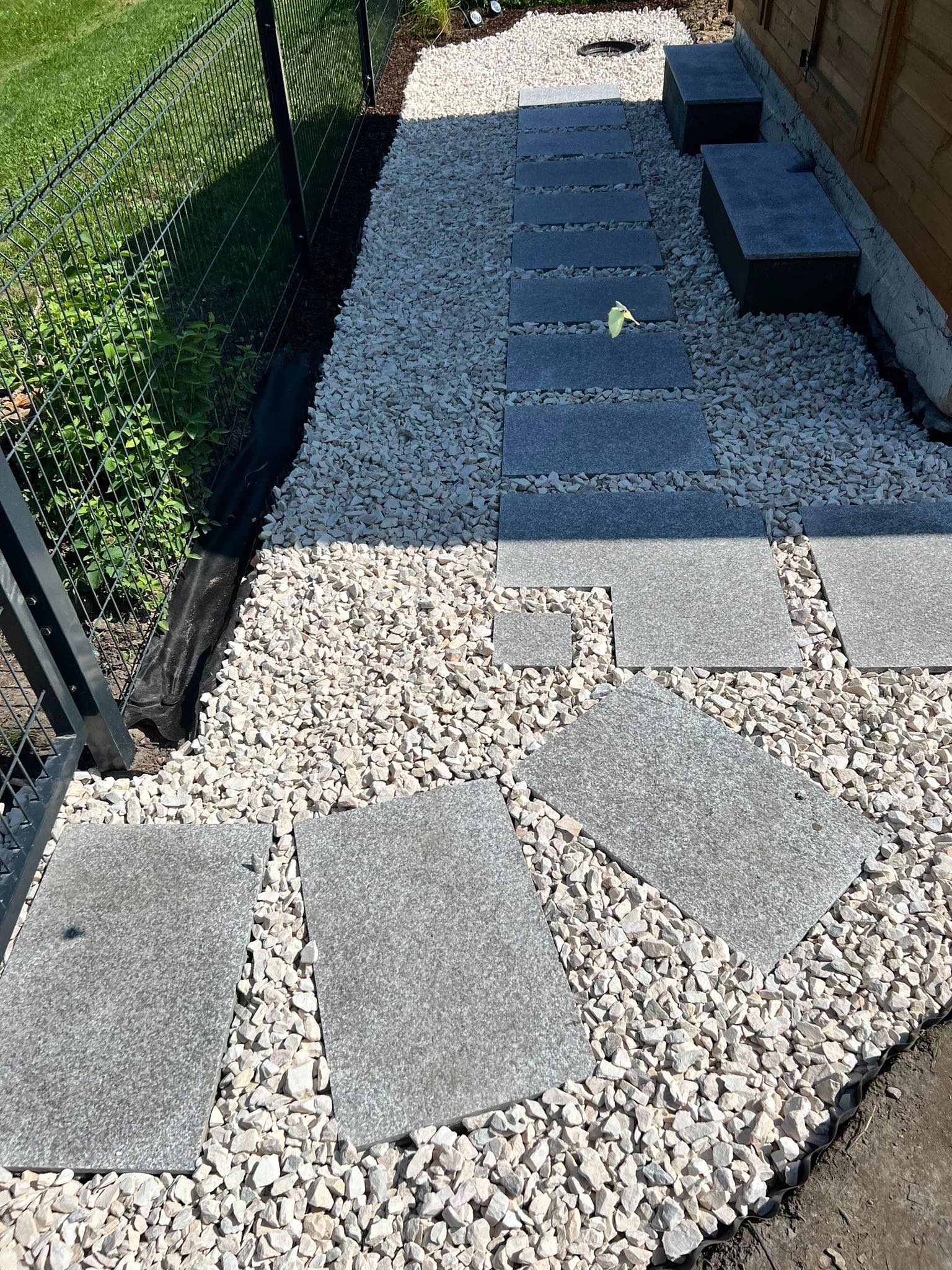 Ścieżka z szarych płyt kamiennych ułożona w białym żwirze, prowadząca do drewnianego budynku z dwoma kamiennymi stopniami, obok czarny płot z siatki.